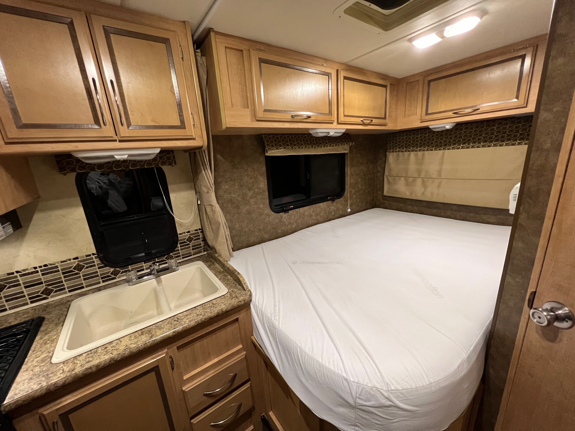 Compact rv rental interior featuring a practical kitchen with sink, a cozy bed, and overhead wood cabinets.. Thor Four Winds 2013