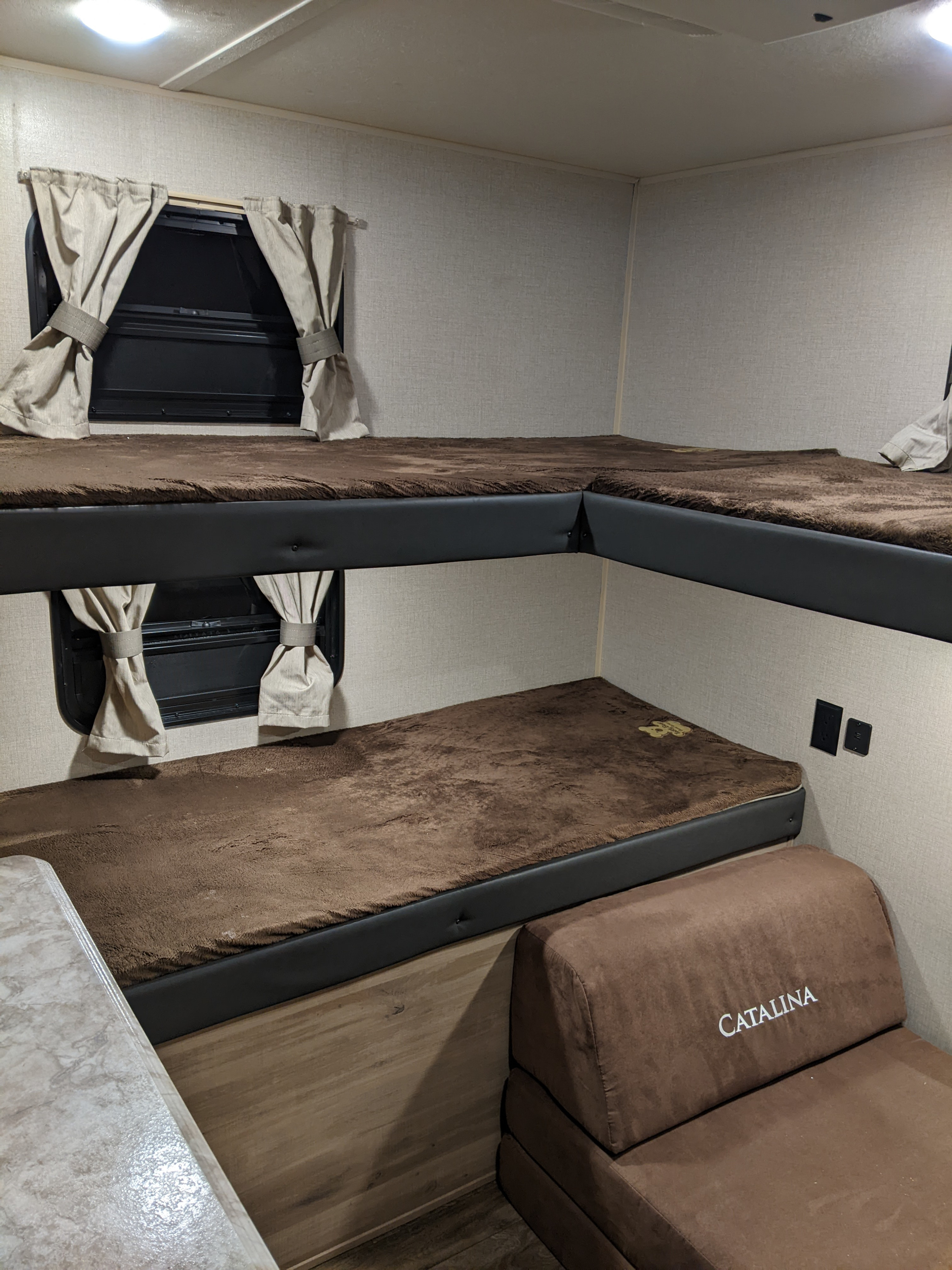Interior of an RV rental: L-shaped bunk beds with cozy brown mattresses, tie-back curtains, and a CATALINA sofa.. Coachmen Catalina Legacy Edition 2020