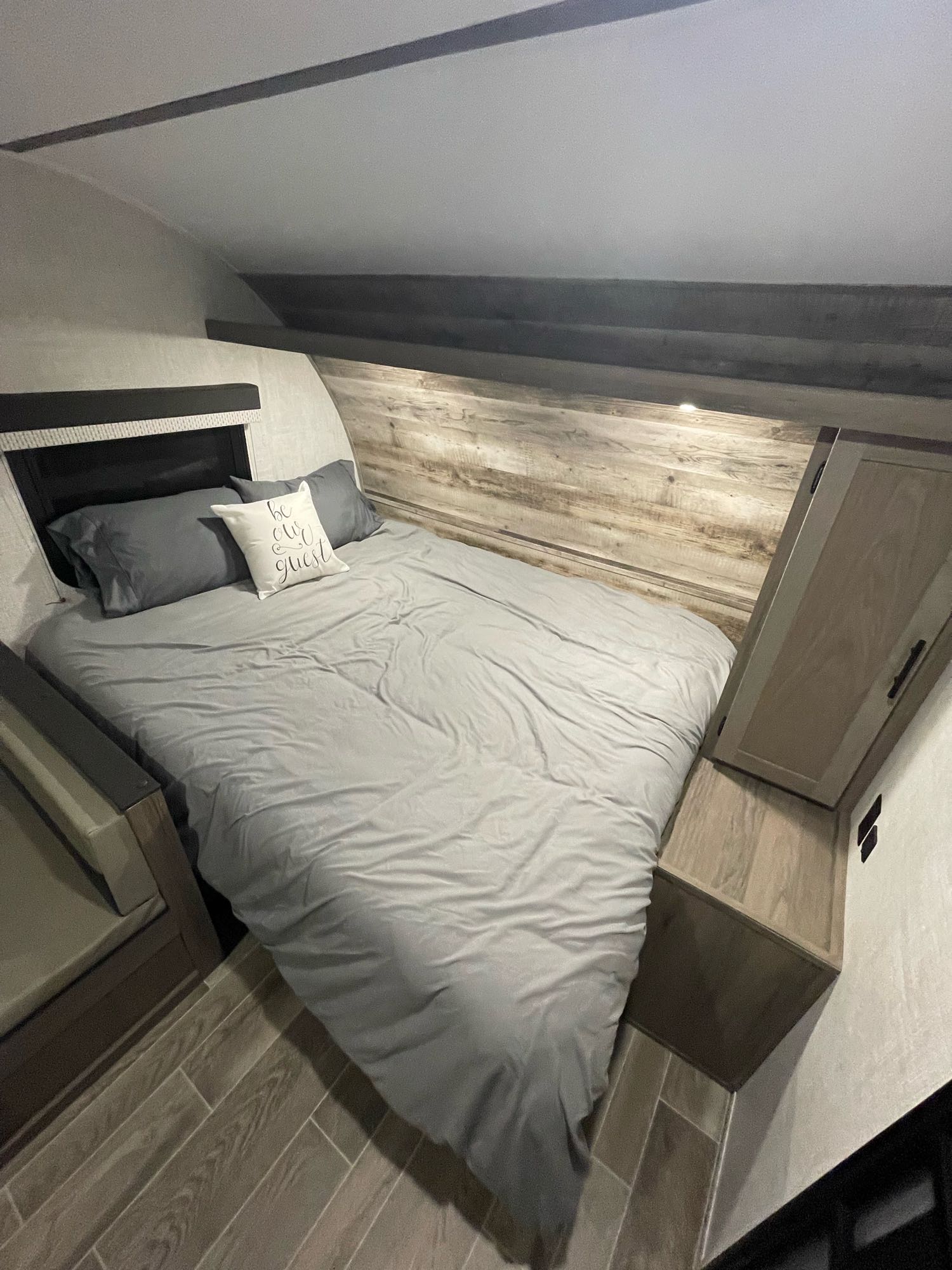 Comfortable RV rental bedroom with grey bedding, a "be our guest" pillow, and cozy wooden interior accents.. Gulf Stream Enlighten 18BH 2022