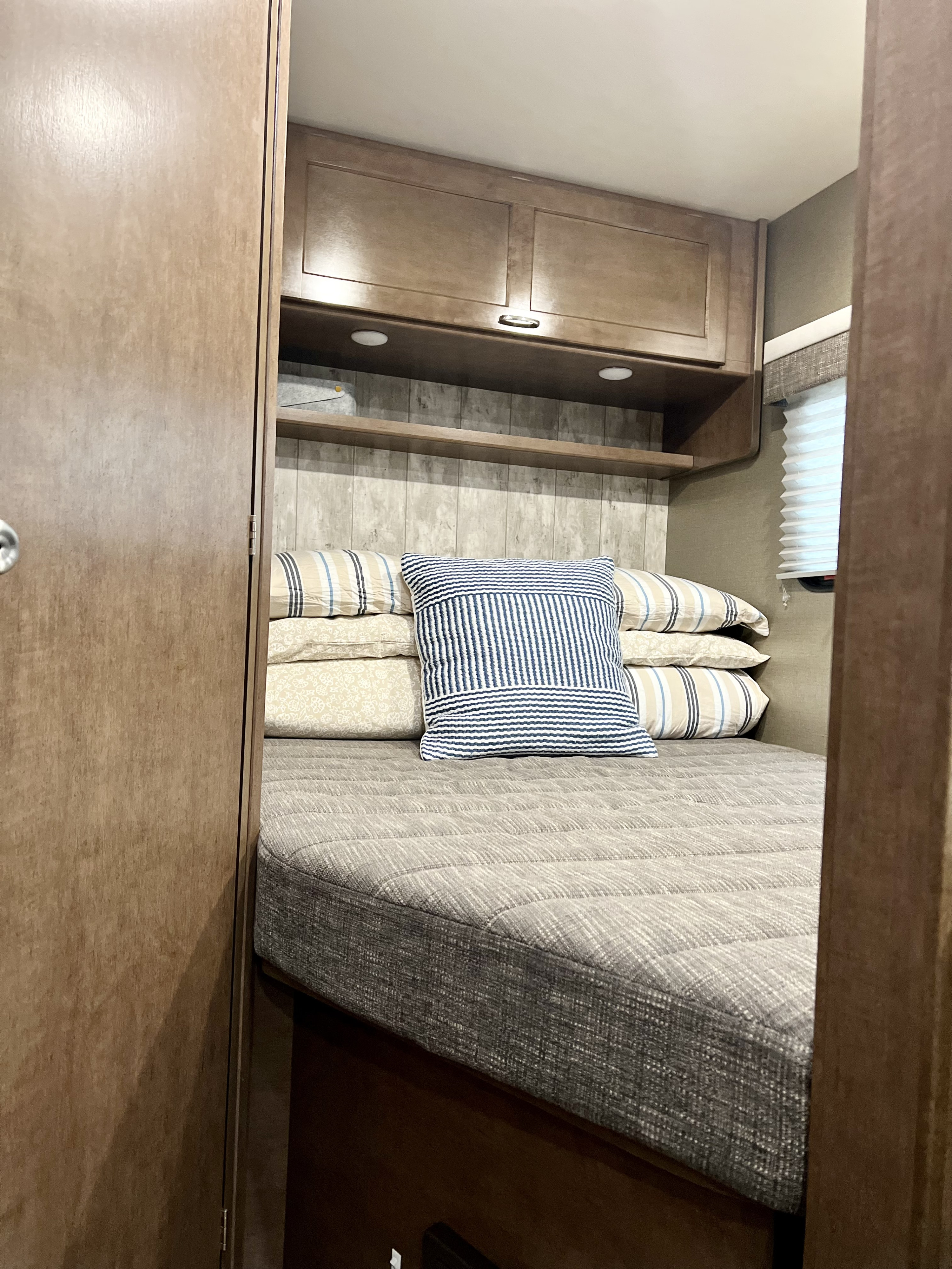 RV rental bedroom interior, showcasing a cozy bed with pillows, overhead cabinets, shelving, and a window. Ideal for compact travel.. Winnebago Outlook 2019