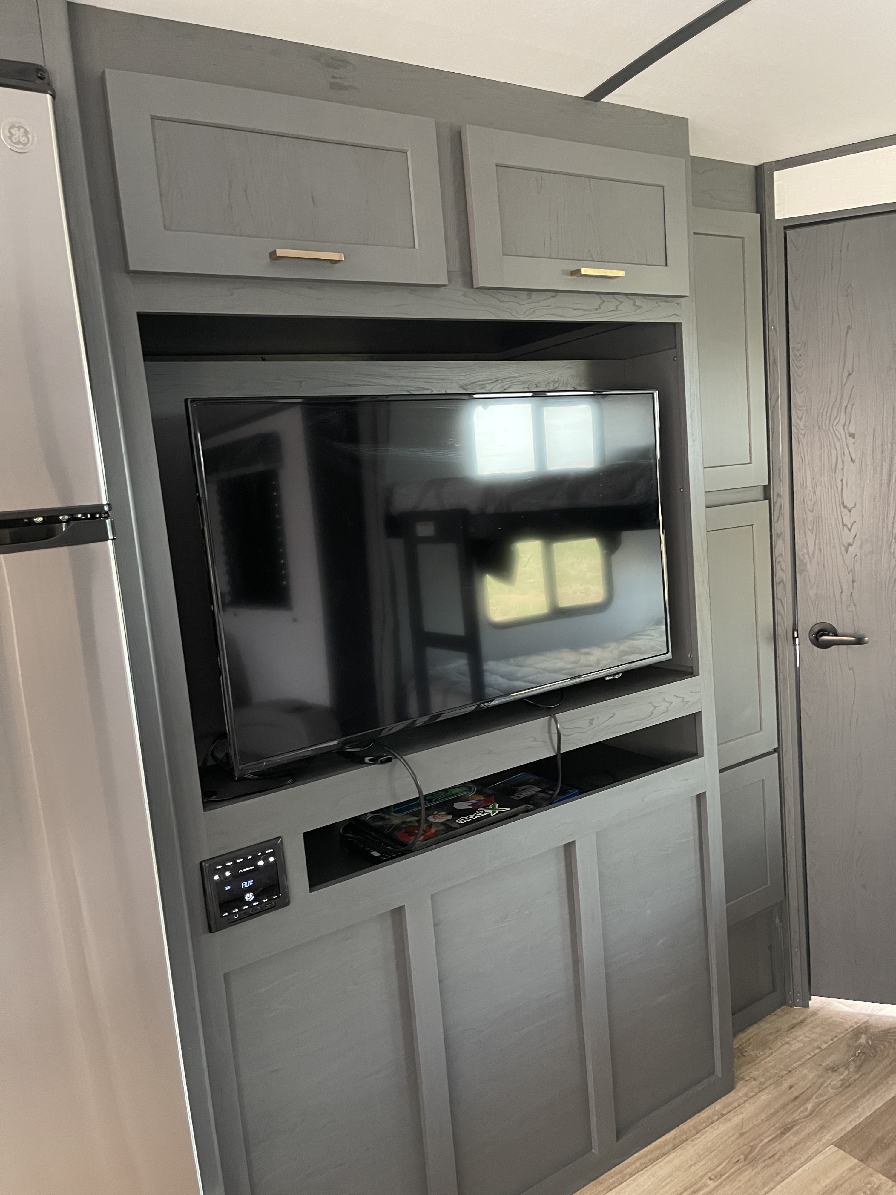 Modern gray RV rental interior with a TV entertainment center, built-in cabinets, and refrigerator. Bunk bed reflection on TV screen.. keystone rv Bullet 2022