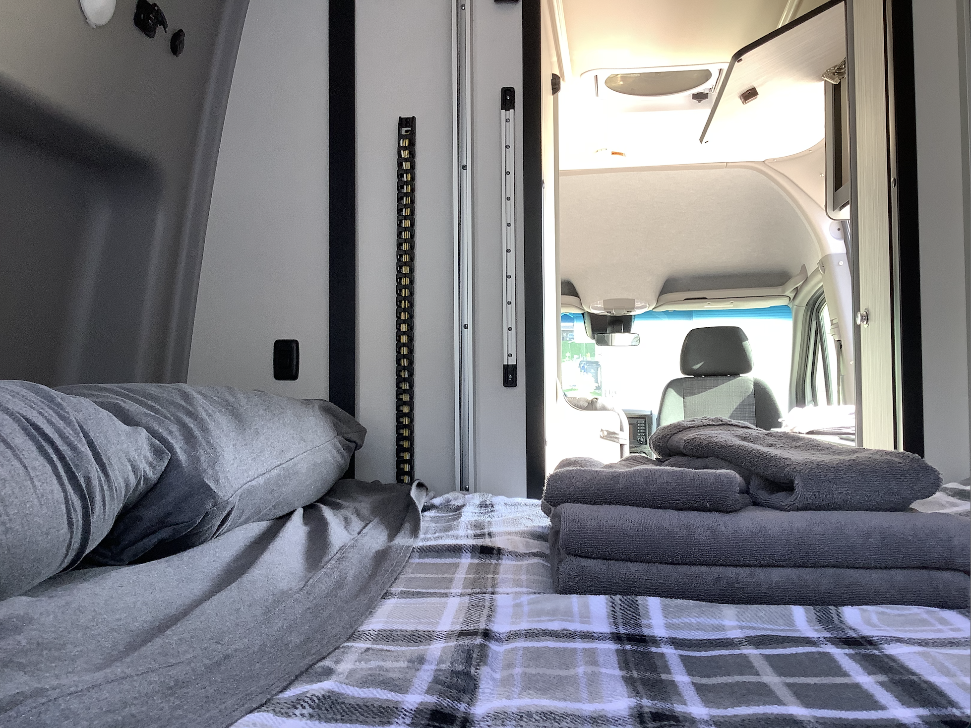 Cozy RV rental interior with a bed, plaid blanket, and stacked towels, looking towards the front cabin.. Winnebago Revel 2019