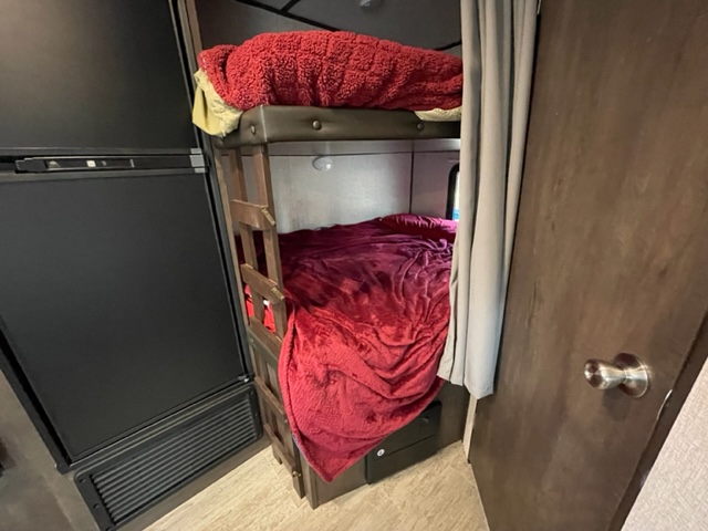 Inside an RV rental: two cozy bunk beds with red blankets, a black refrigerator, and privacy curtain.. Dutchmen Aspen Trail Trailer 2020