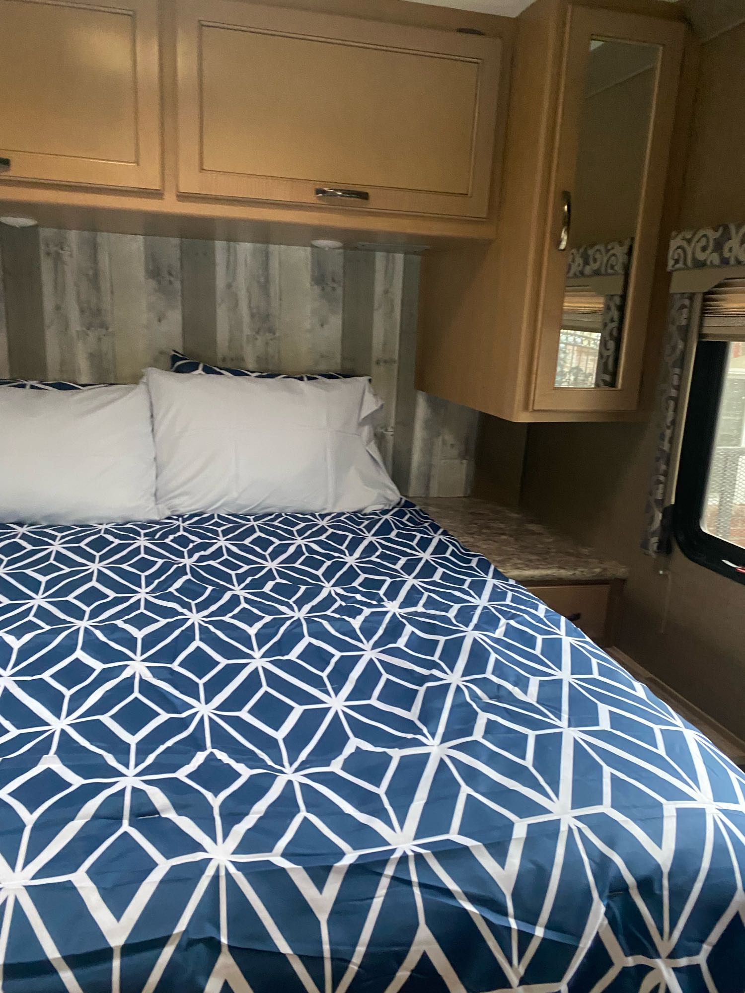 Cozy RV rental bedroom featuring a bed with blue geometric bedding, overhead cabinets, and a window for comfortable travel.. Thor Four Winds 2019