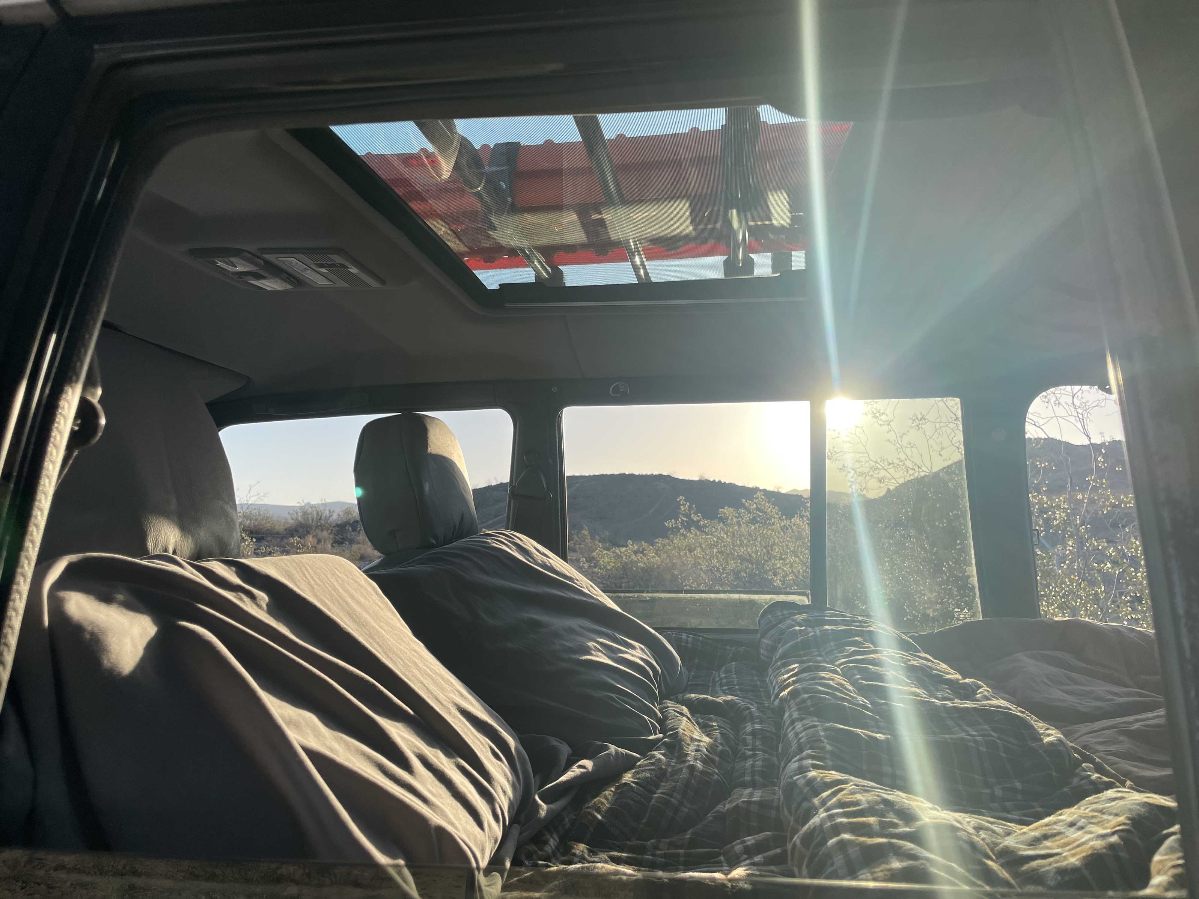 Inside an RV rental camper: cozy bedding with a plaid blanket, bright mountain view, and sunlight through the sunroof.. Custom Camper 1998