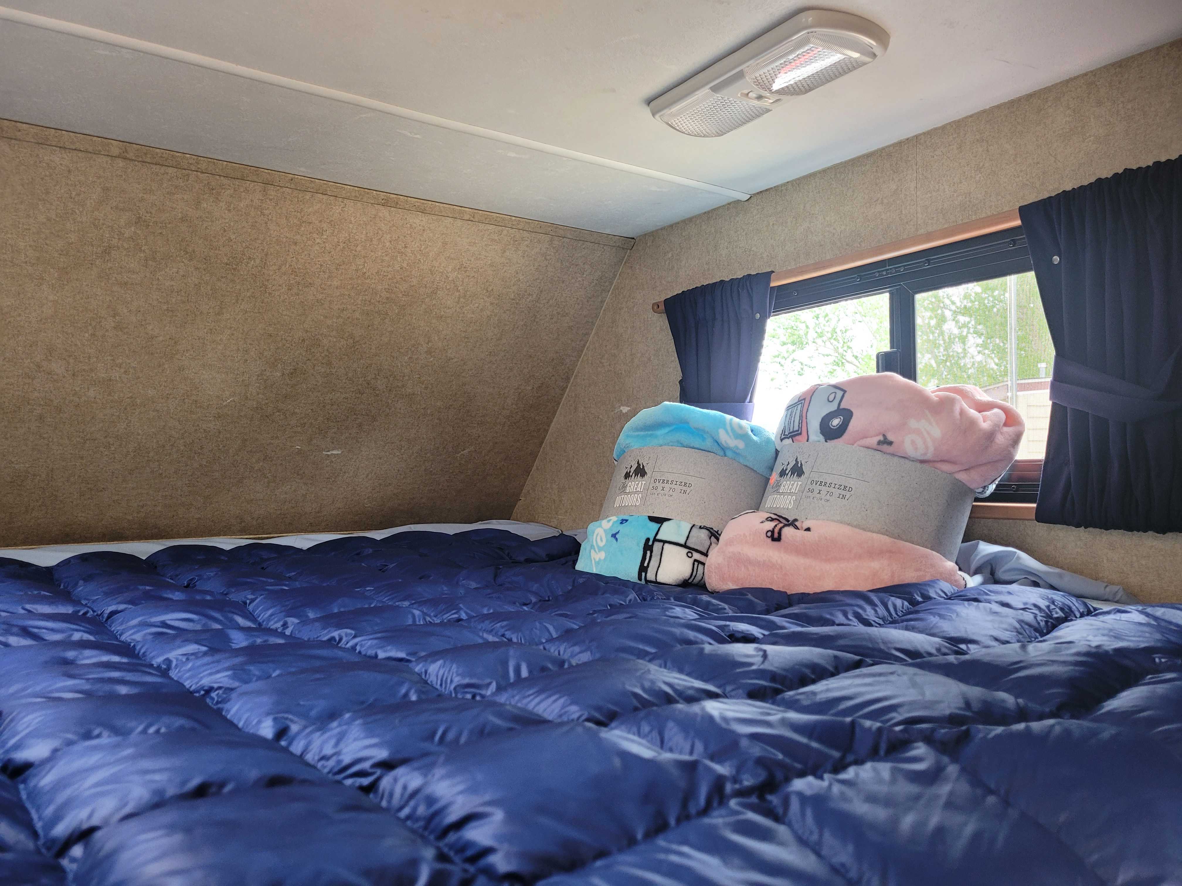Cozy RV rental bed area featuring a blue comforter & "Great Outdoors" blankets, with a window and light.. Thor Majestic 2017