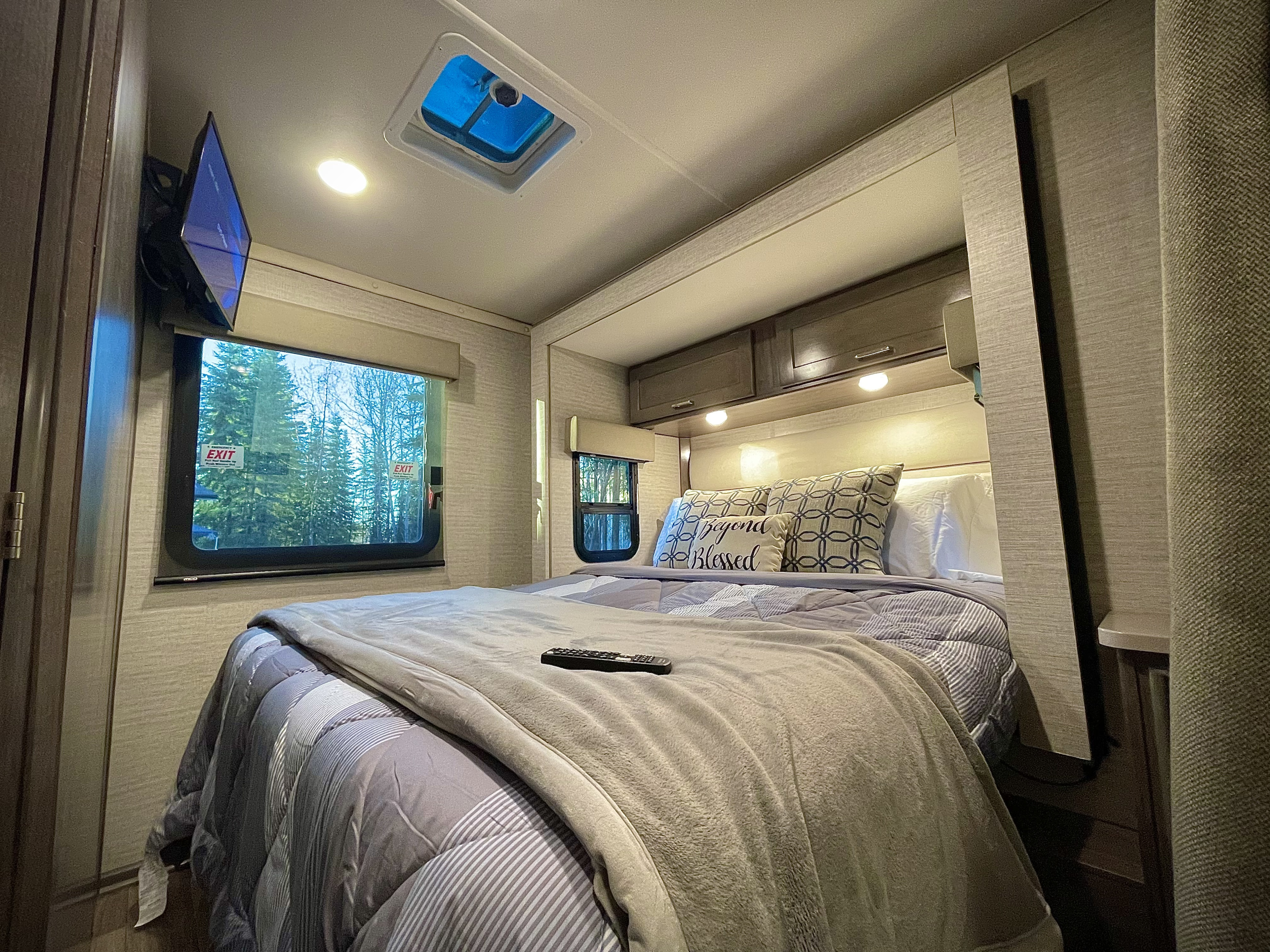 Cozy RV rental bedroom featuring a comfortable bed with gray bedding, a wall-mounted TV, and a window overlooking trees.. Winnebago Minnie Winnie 2020