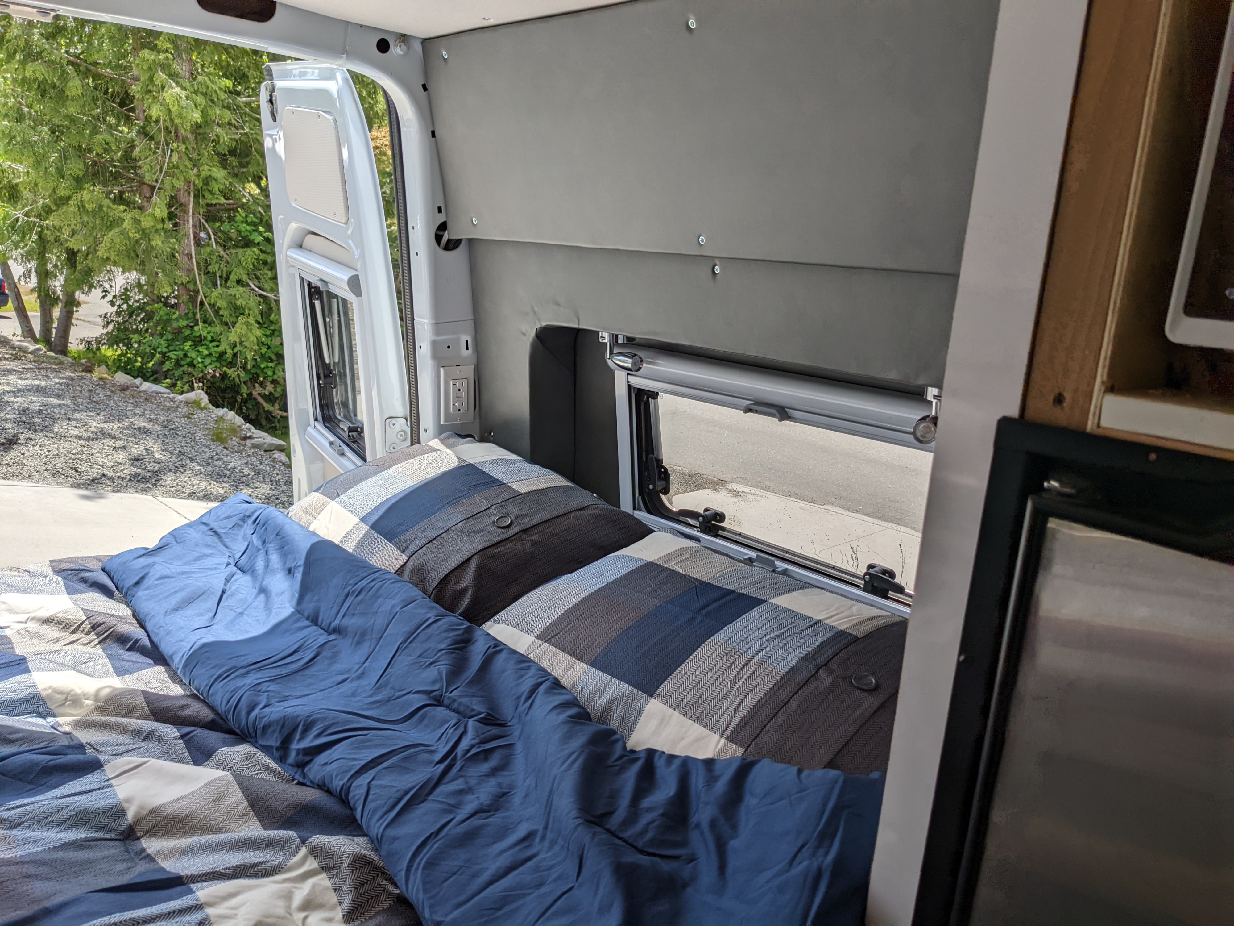 Cozy RV rental interior with a comfy bed, plaid bedding, and an open window offering a view of lush trees.. Other Sprinter 2016