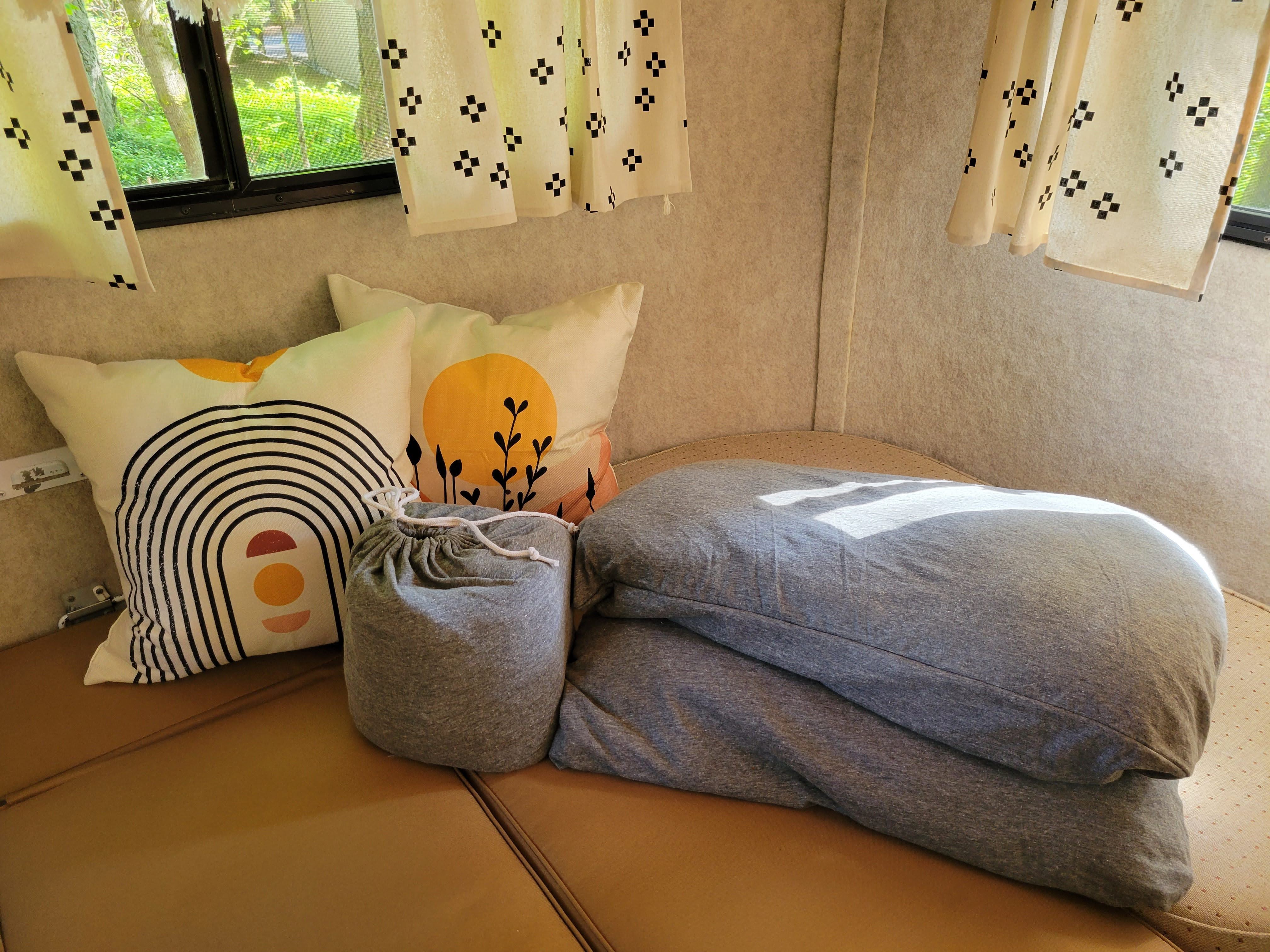 Bright RV rental interior with patterned white curtains, colorful throw pillows, and folded grey bedding on brown cushions.. Scamp 16' 1992