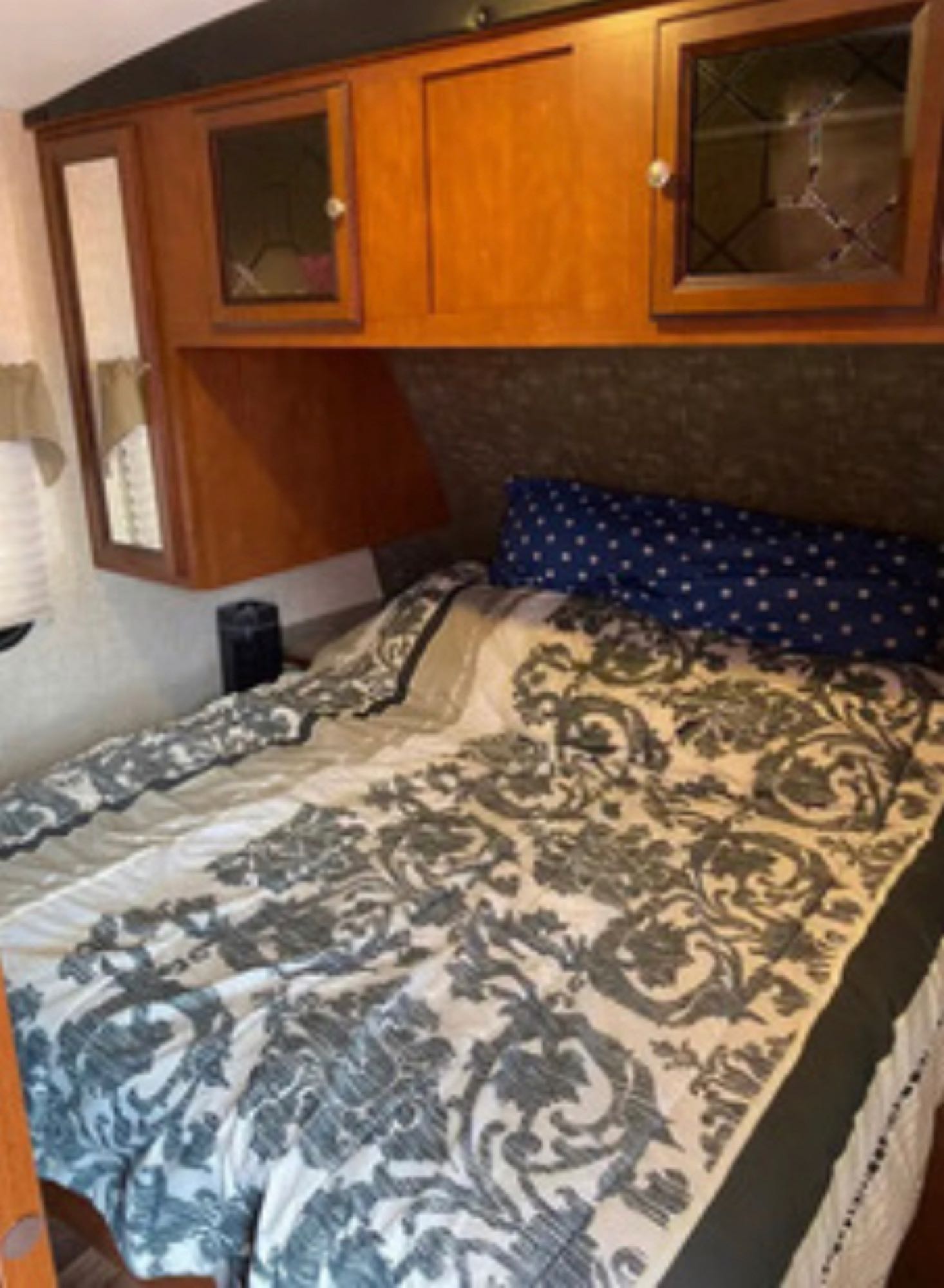 This RV rental features a cozy bedroom with patterned bedding, overhead storage cabinets, and a side mirror.. Heartland RVs Mallard 2016