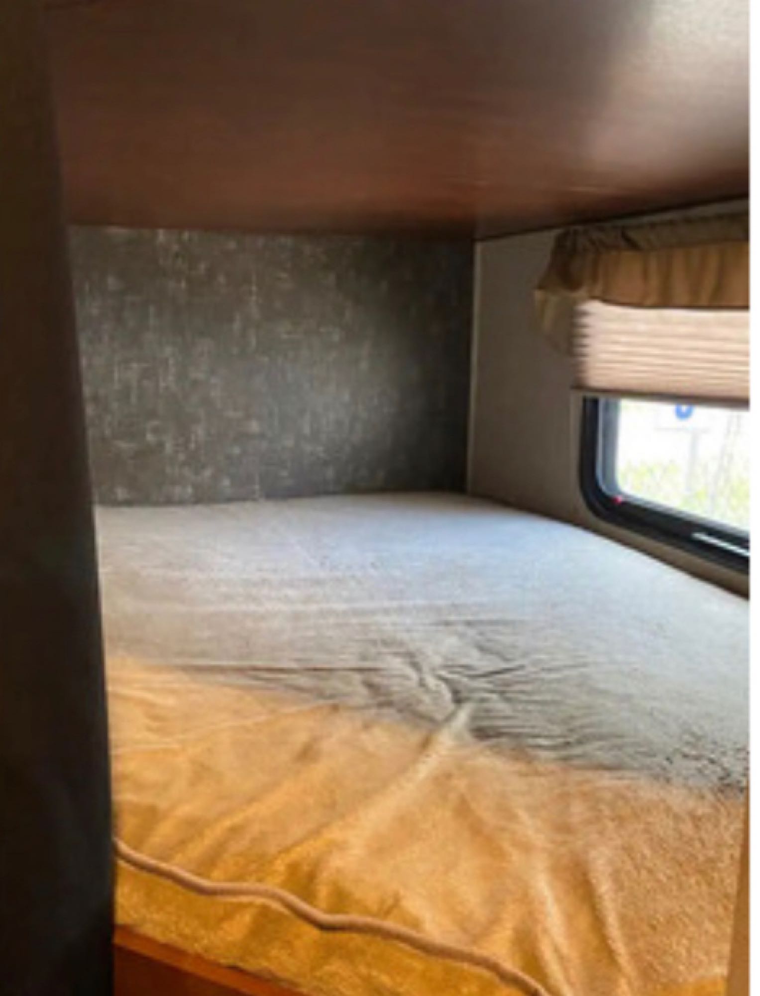 View of a compact bed in an RV rental, featuring a textured wall, window with blinds, and soft blanket.. Heartland RVs Mallard 2016