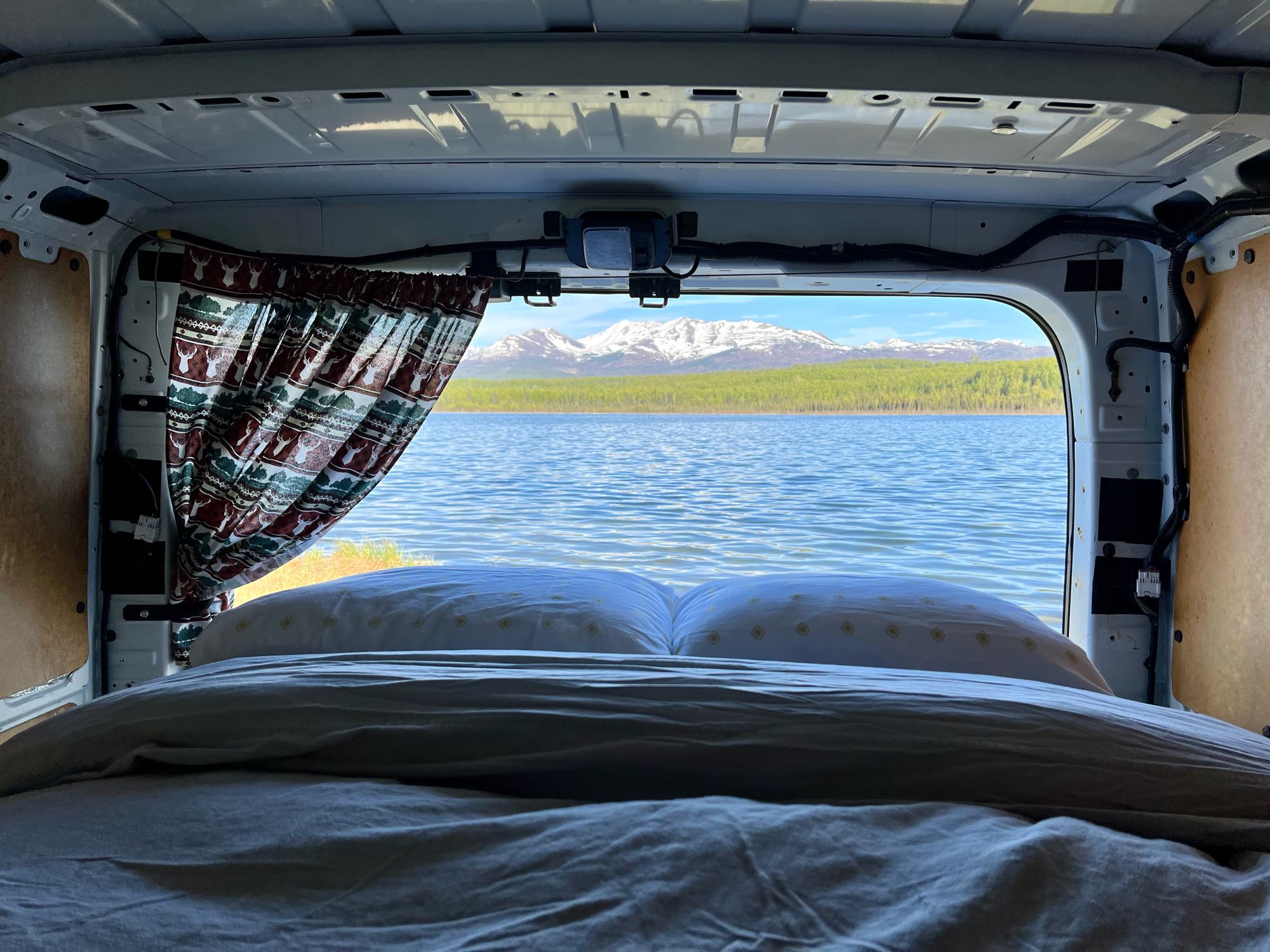 Scenic RV rental view: Cozy bed overlooking a calm lake and snow-capped mountains. Perfect for van life adventures!. Nissan Other 2015