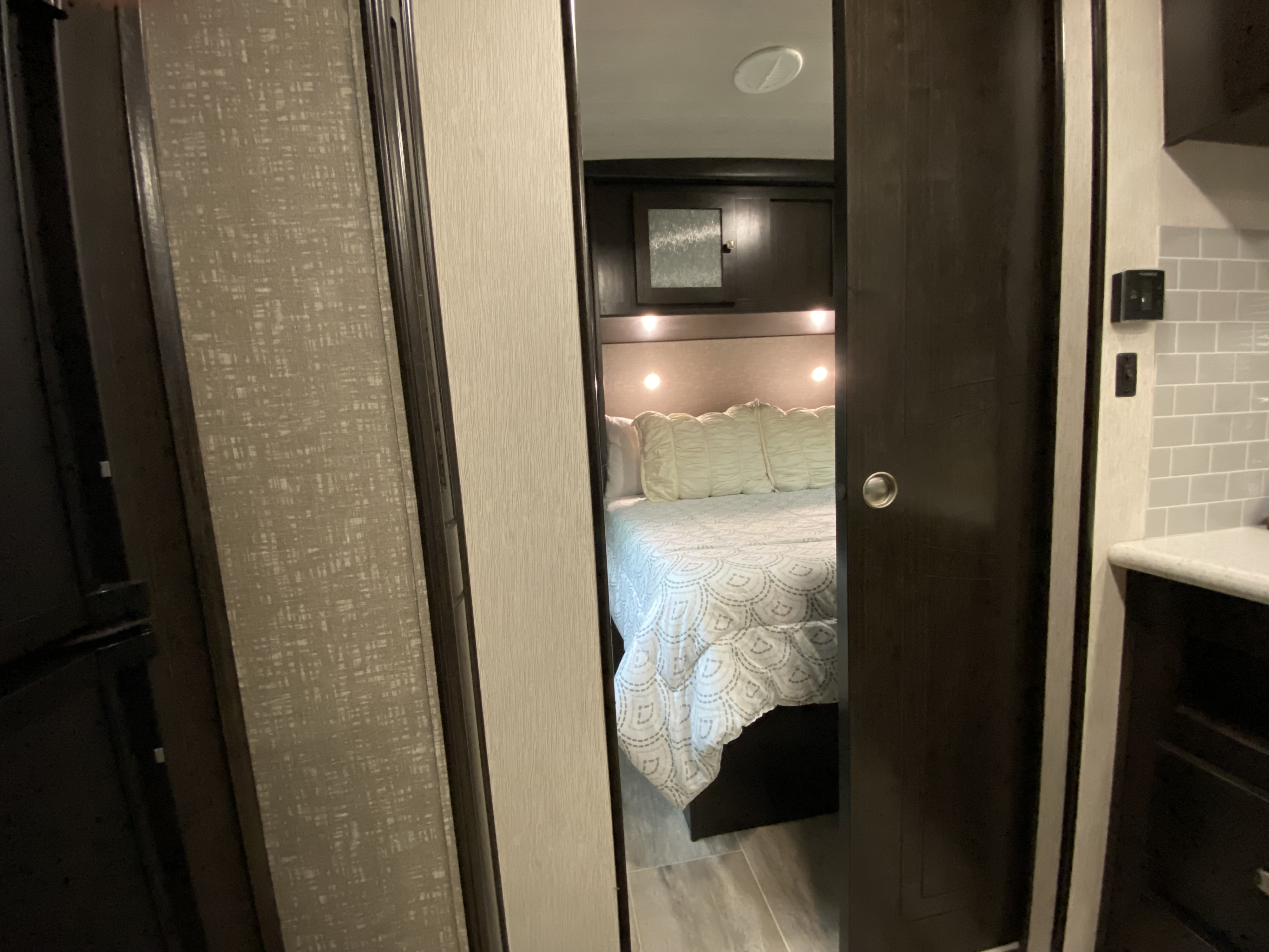 Inside an rv rental, a bedroom with a cozy bed and headboard lights is visible through a doorway, with textured walls and kitchen.. Heartland RVs North Trail 2019