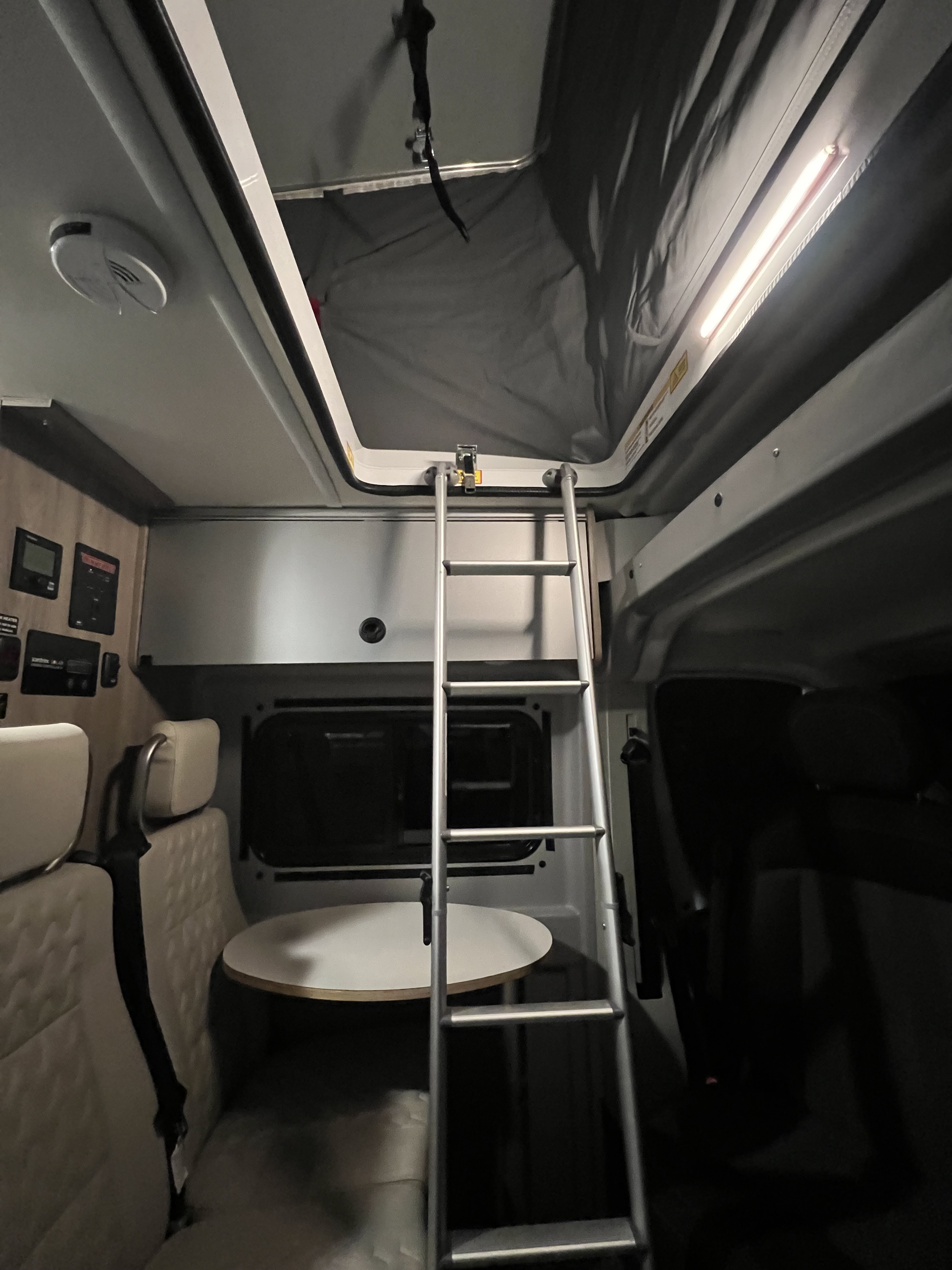 Inside an RV rental: An illuminated ladder leads to a pop-top roof tent, with a table, seating, and control panel visible.. Winnebago Solis 2022