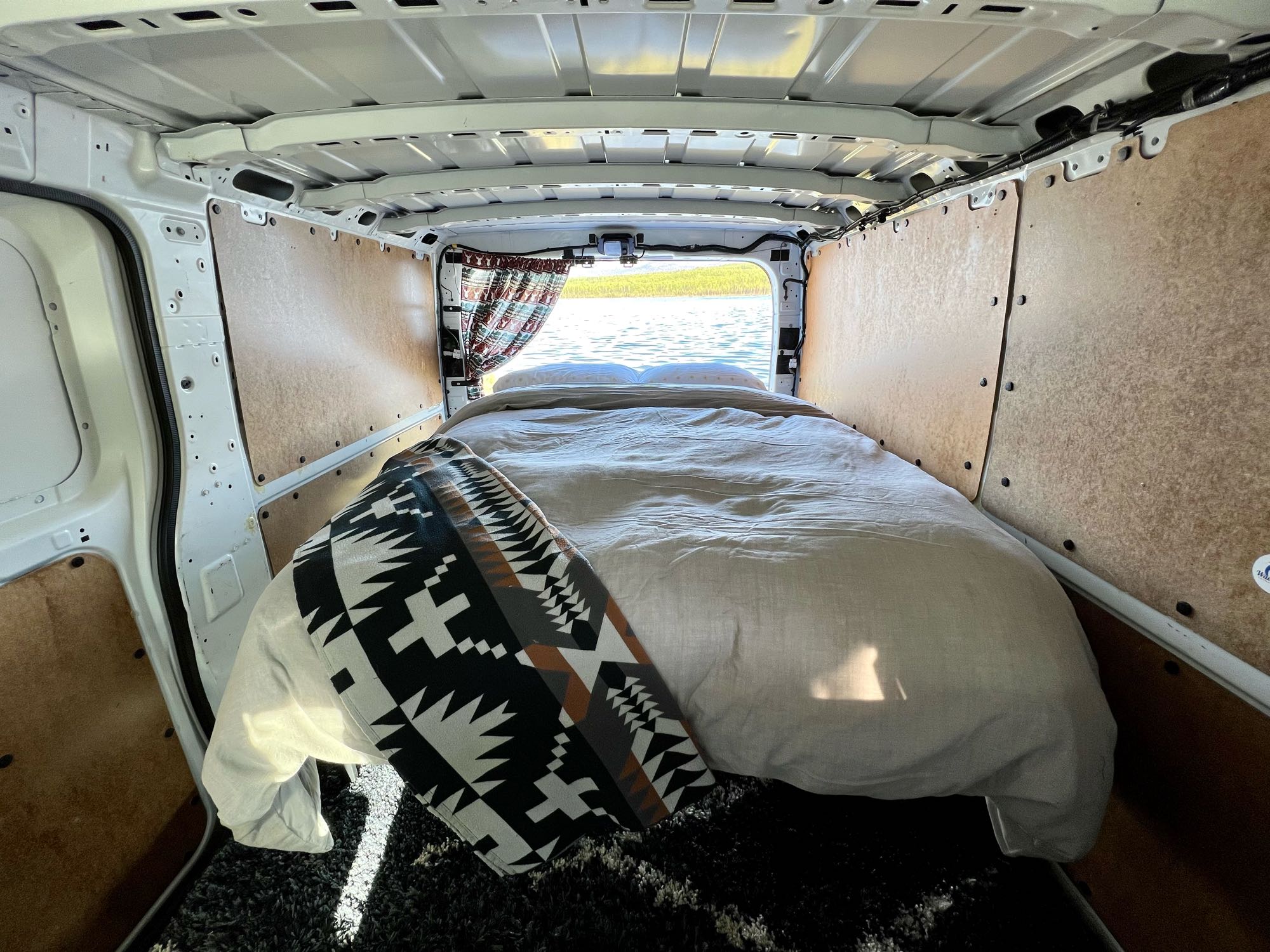 RV rental interior: A made bed with a patterned blanket looks out to a lake view through open rear doors.. Nissan Other 2015