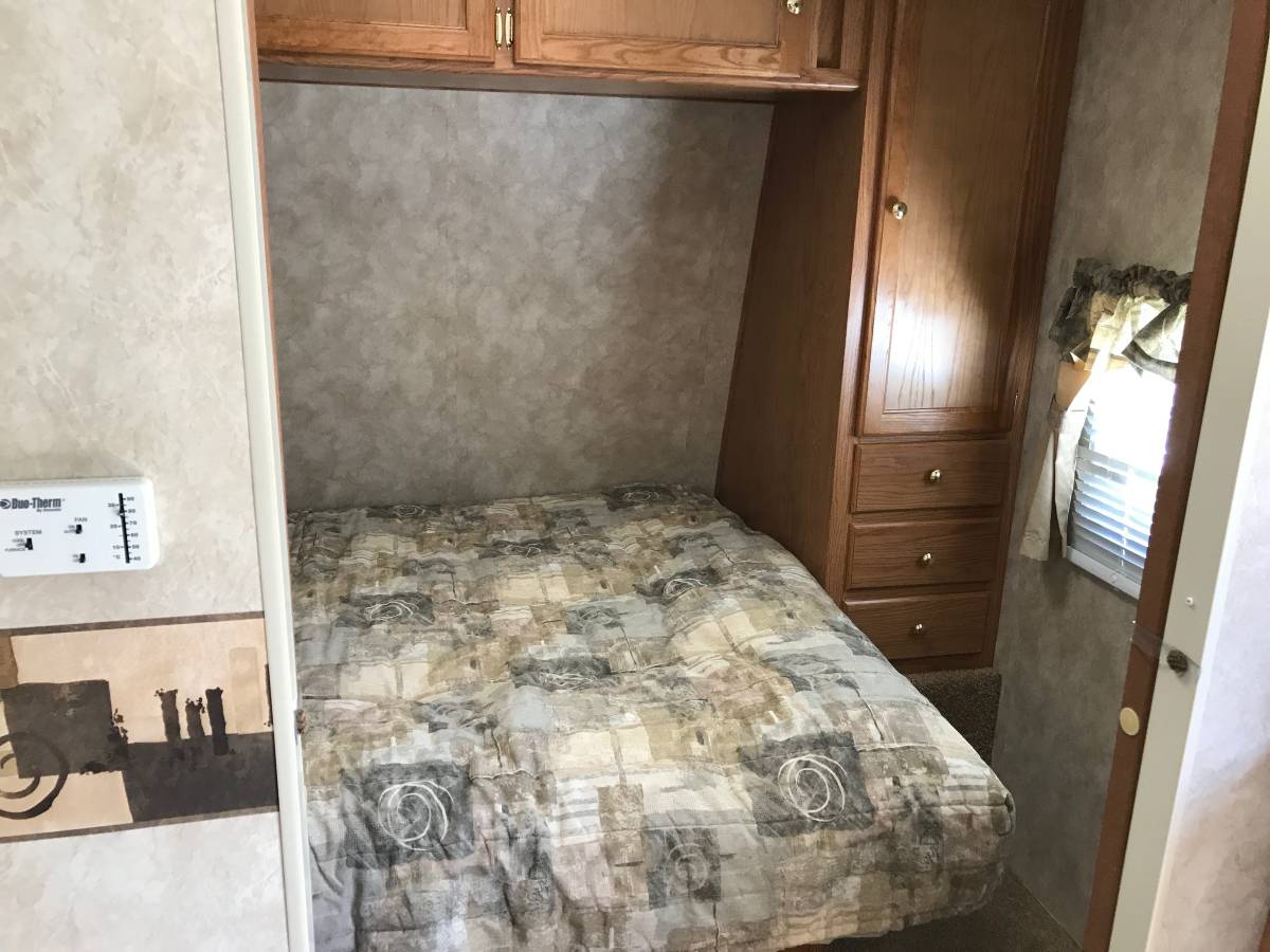 View of a compact RV rental bedroom with a patterned bed, wooden storage cabinet, and wall-mounted thermostat.. Keystone RV Springdale 2006