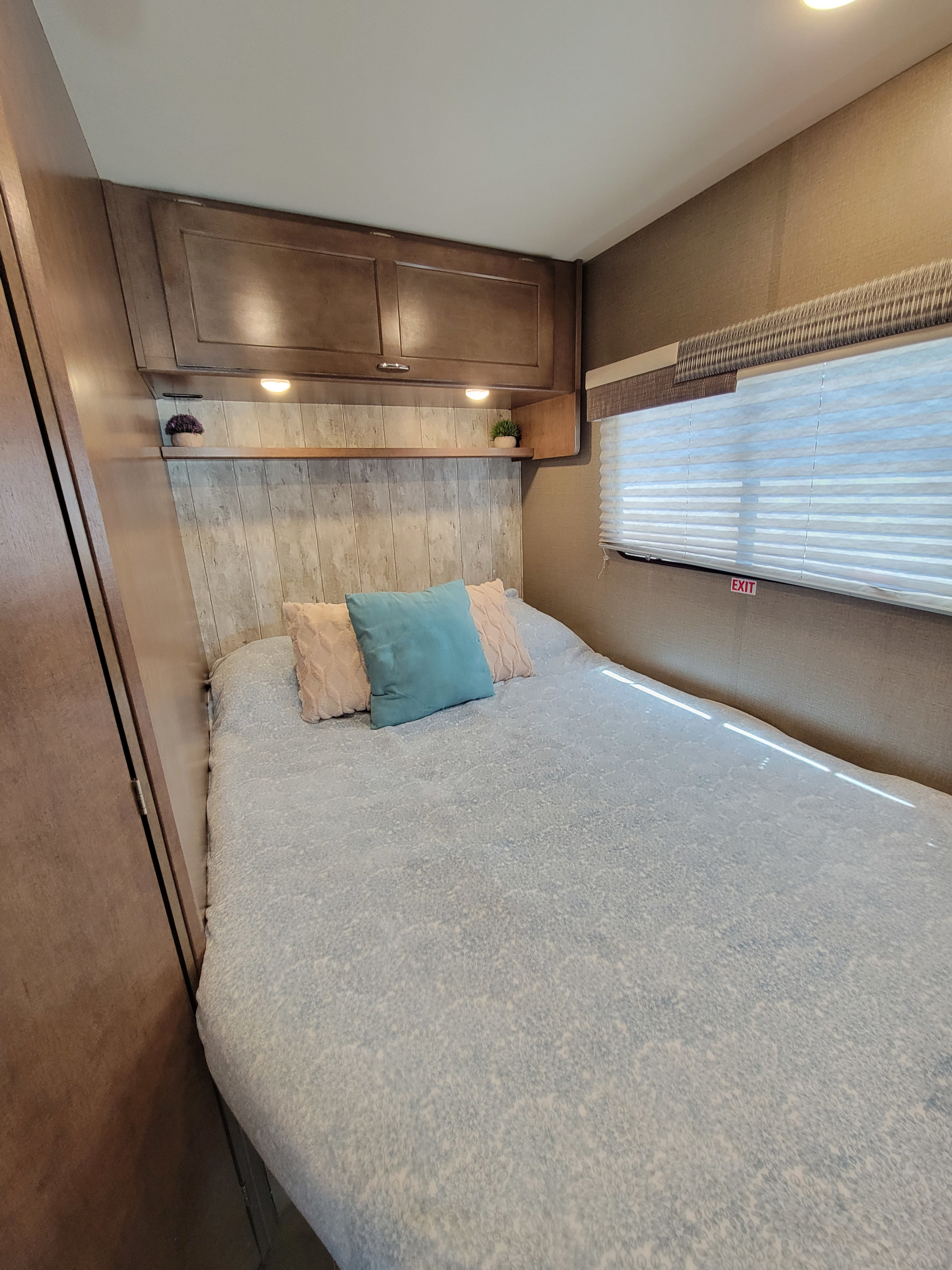 Cozy RV rental bedroom with a comfortable bed, accent pillows, overhead cabinets, and a window with blinds.. Winnebago Outlook 2019