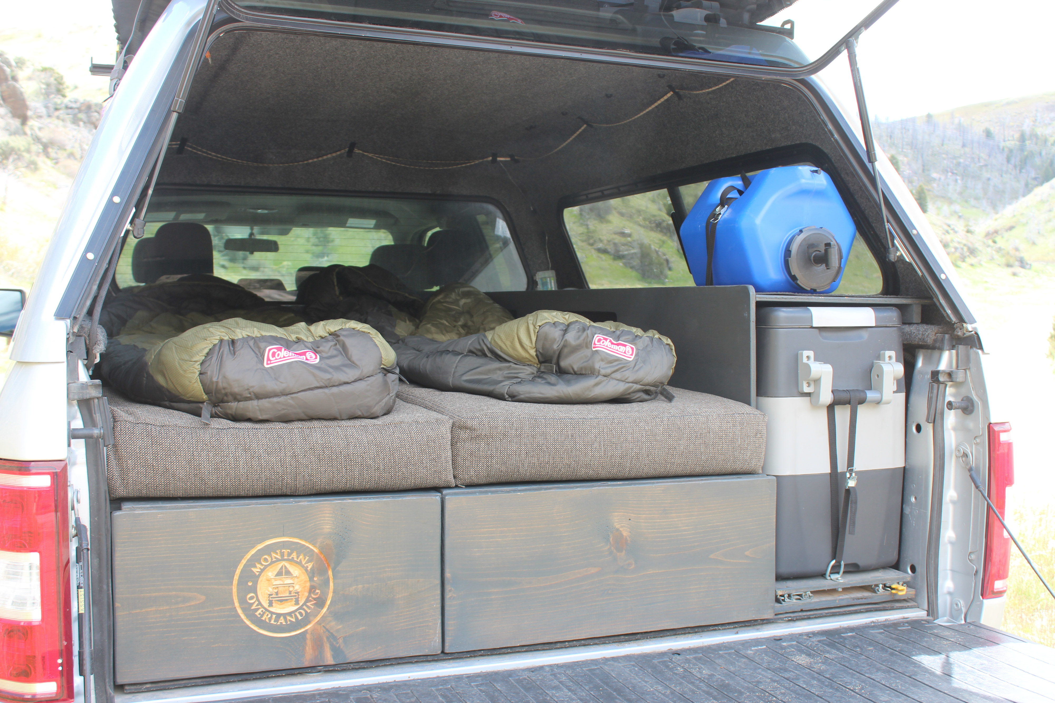 RV rental truck bed camper setup: features a comfortable sleeping area with bags, storage drawers, fridge, and water for overlanding.. Ford Ford F150 2018