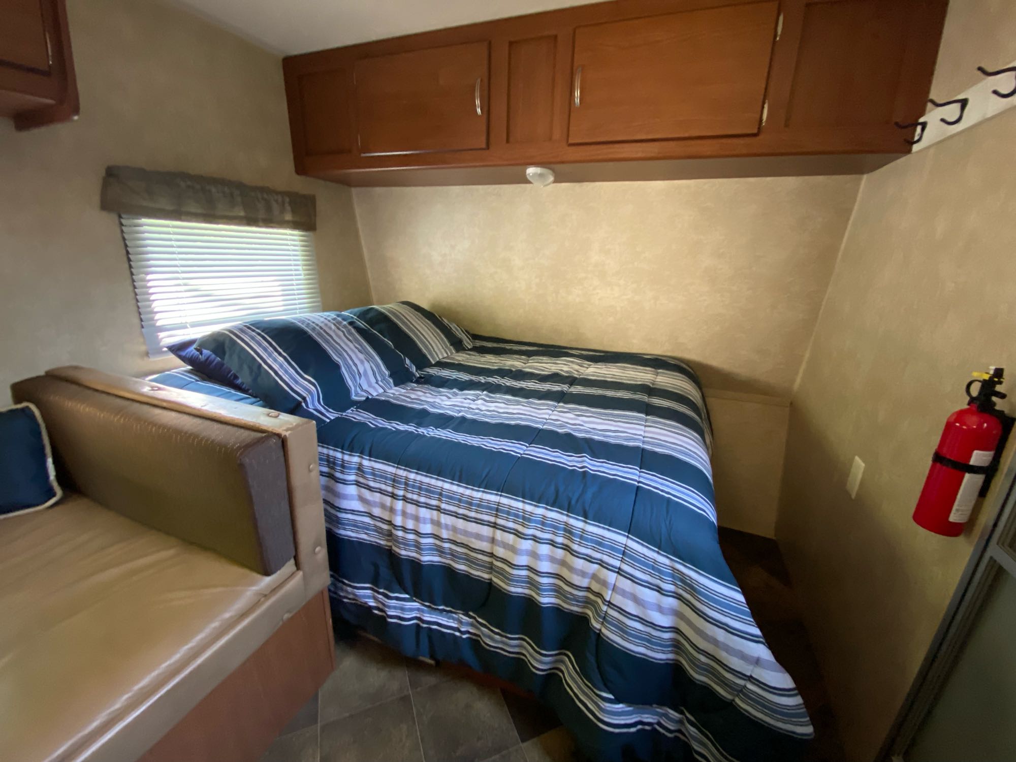 RV rental interior: A compact bedroom with striped bed, overhead cabinets, window, and a wall-mounted fire extinguisher.. Forest River Salem Cruise Lite 2015