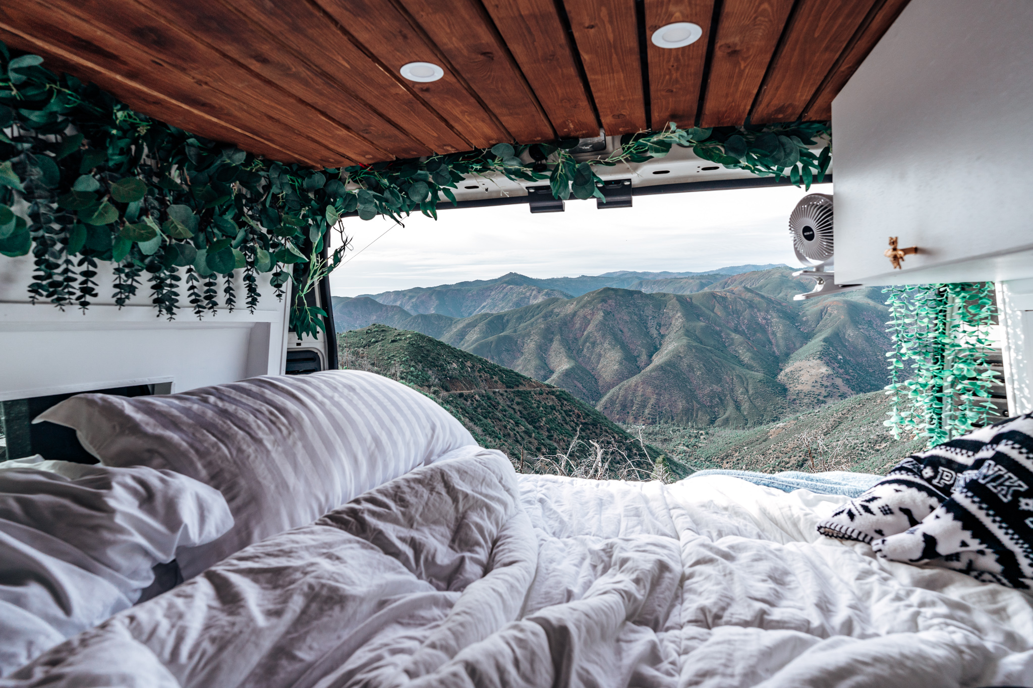 Cozy RV rental interior: a comfortable bed with white linens, leafy green decor, and an expansive mountain vista.. RAM Promaster 2015