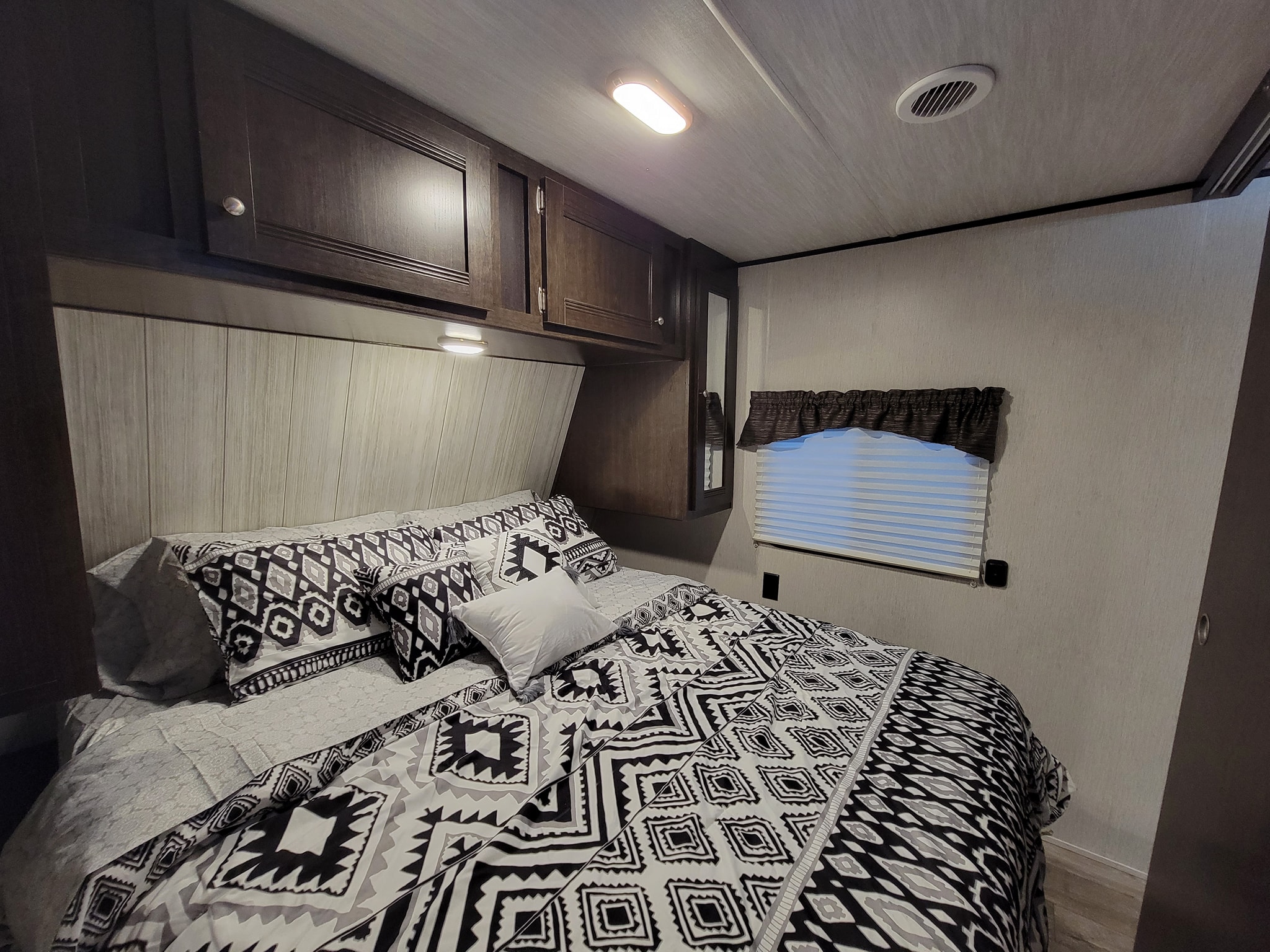 Spacious RV rental bedroom with modern black/white bedding, overhead cabinets, soft lighting, and a window blind.. Heartland RVs Pioneer 2019