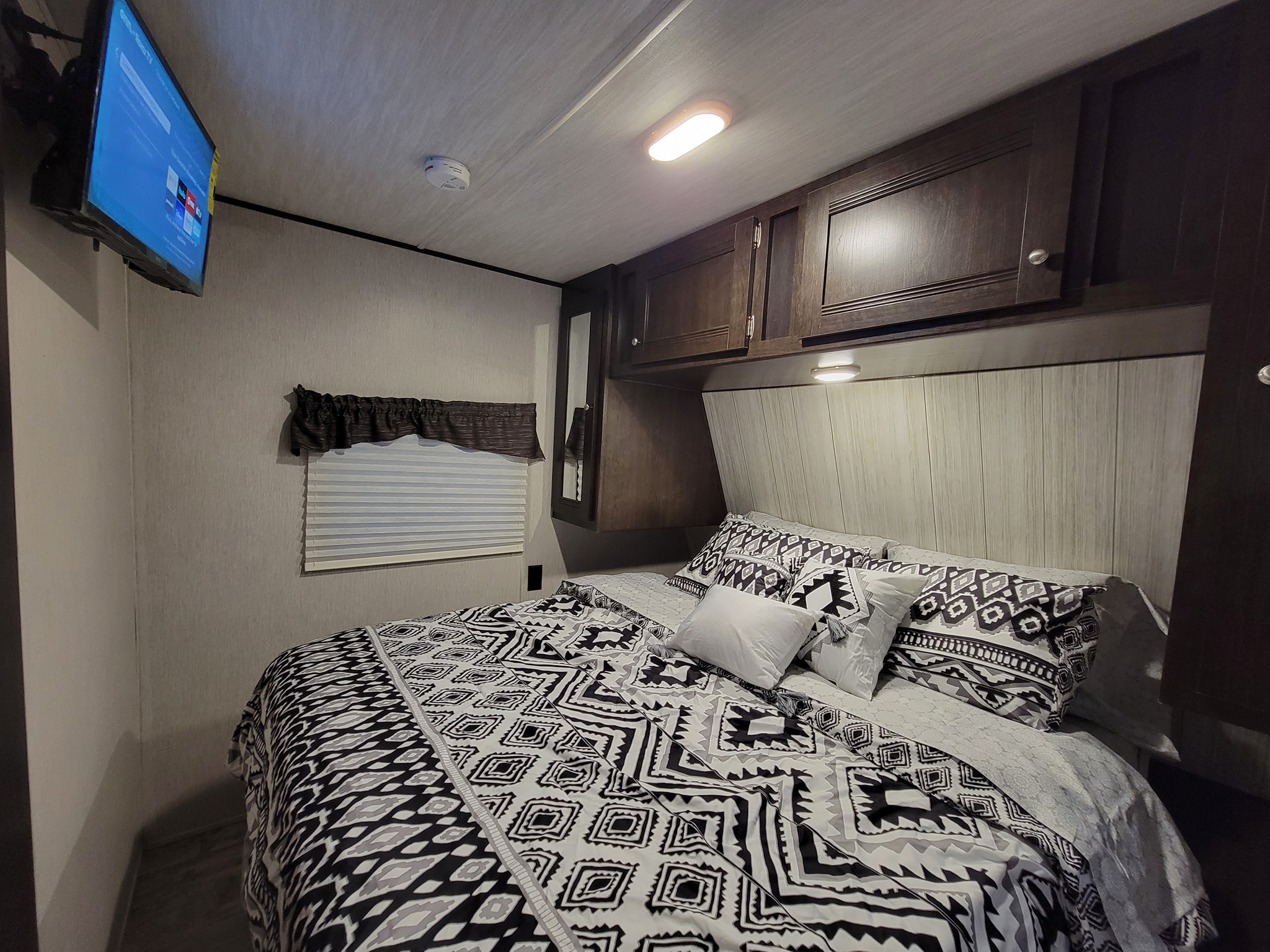 RV rental bedroom: Interior shows a bed with black/white geometric bedding, a wall-mounted TV, and dark wood cabinets.. Heartland RVs Pioneer 2019