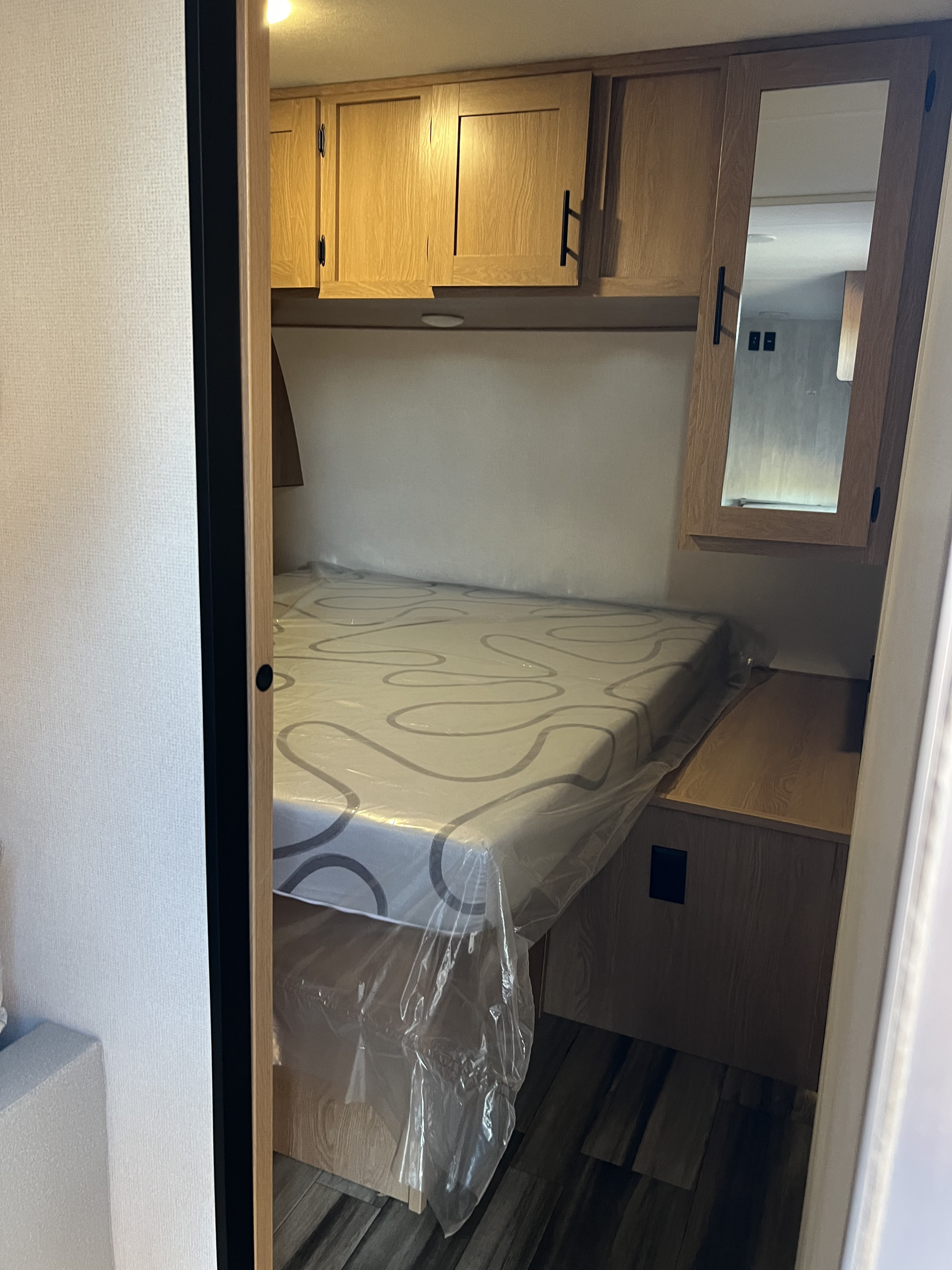 Inside an RV rental, a compact bedroom features a bed (still in plastic), wood cabinets, and a mirrored wardrobe door.. Avenger other 2022