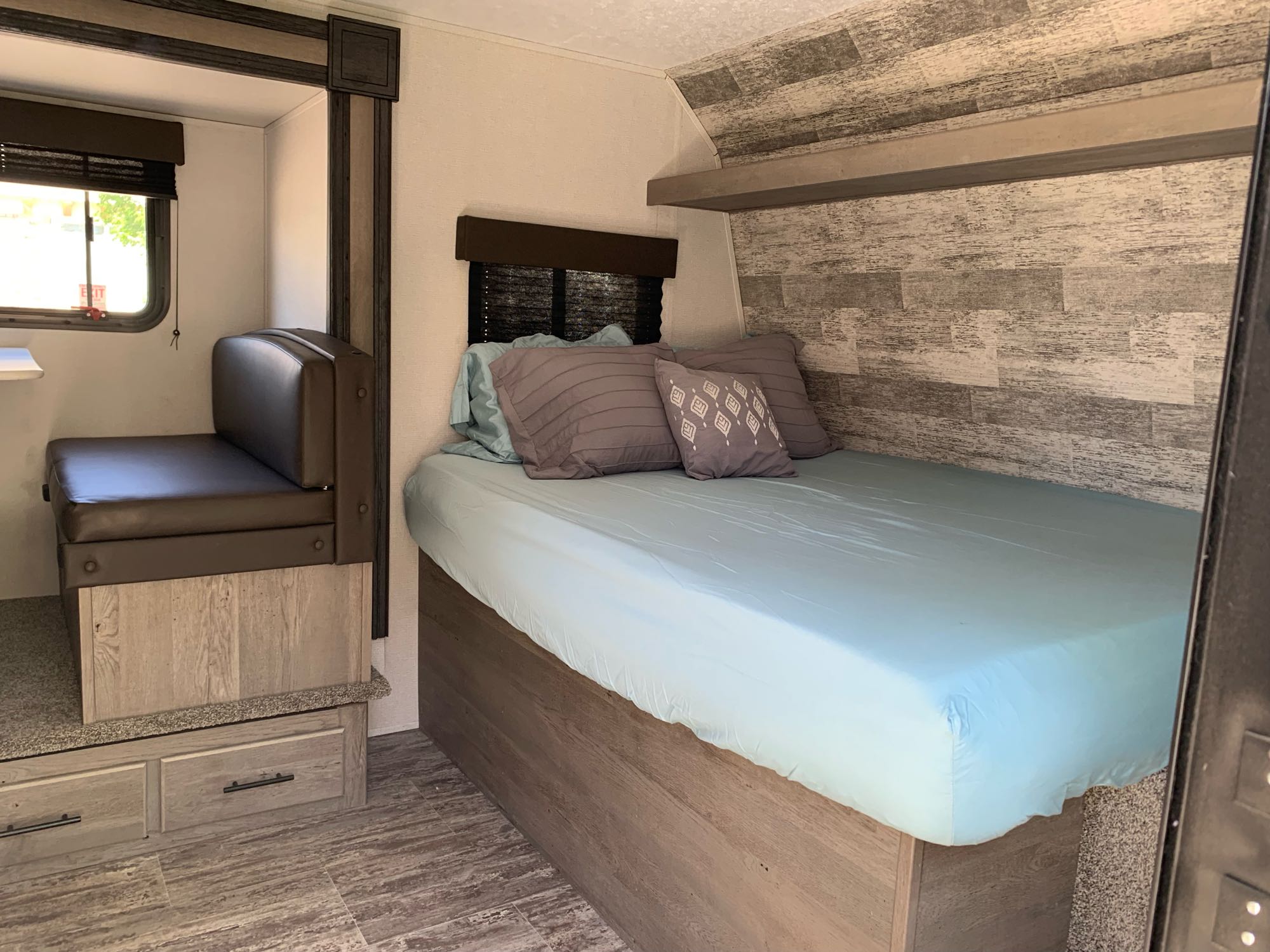 Inside an RV rental: view of a compact bedroom with bed & pillows, adjacent to a seating area with a window & storage drawers.. Springdale Bunkhouse 2021
