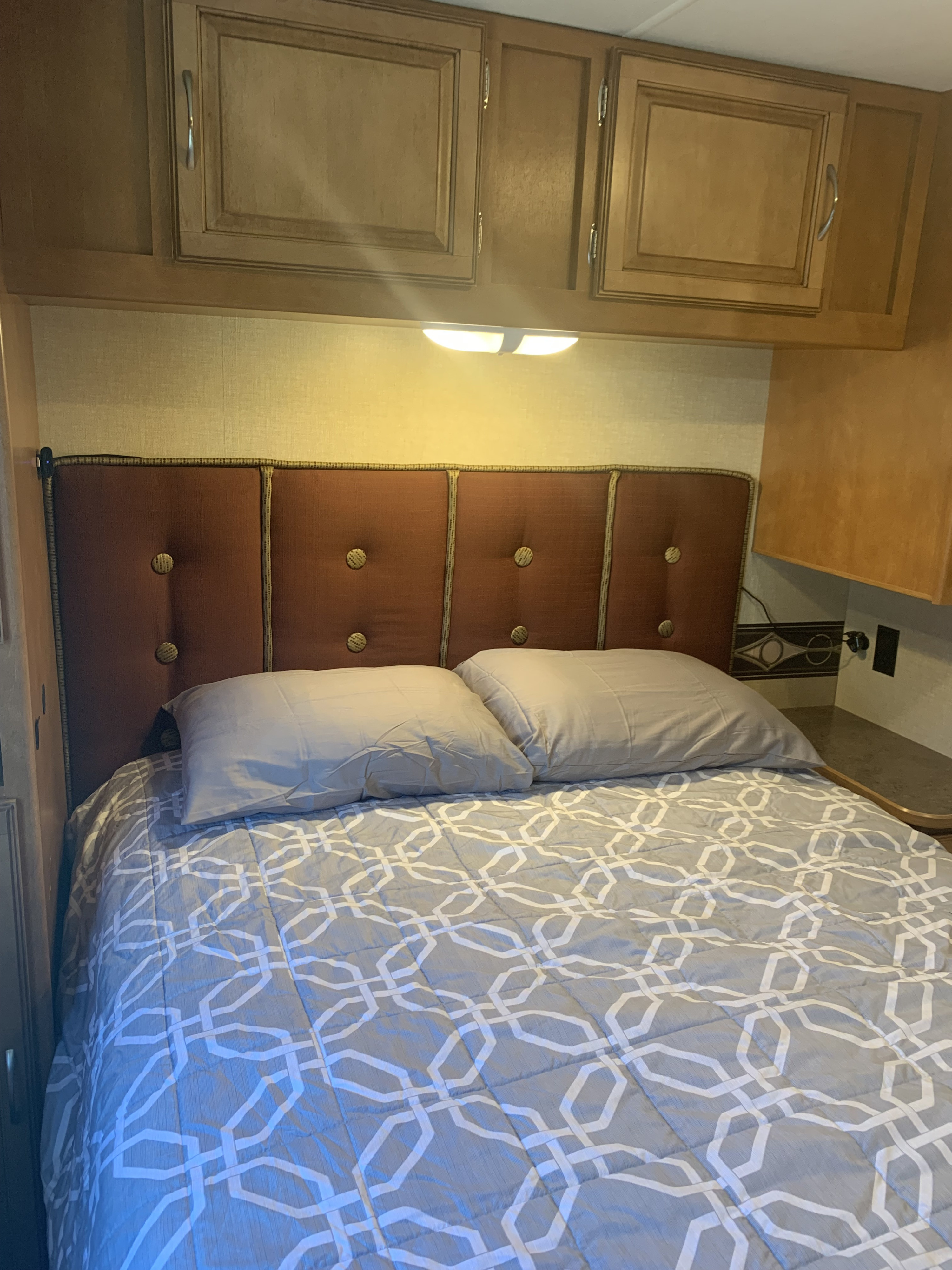 An RV rental bedroom featuring a comfy bed with a grey geometric comforter, padded headboard, and overhead cabinets.. Winnebago Minnie Winnie 2016
