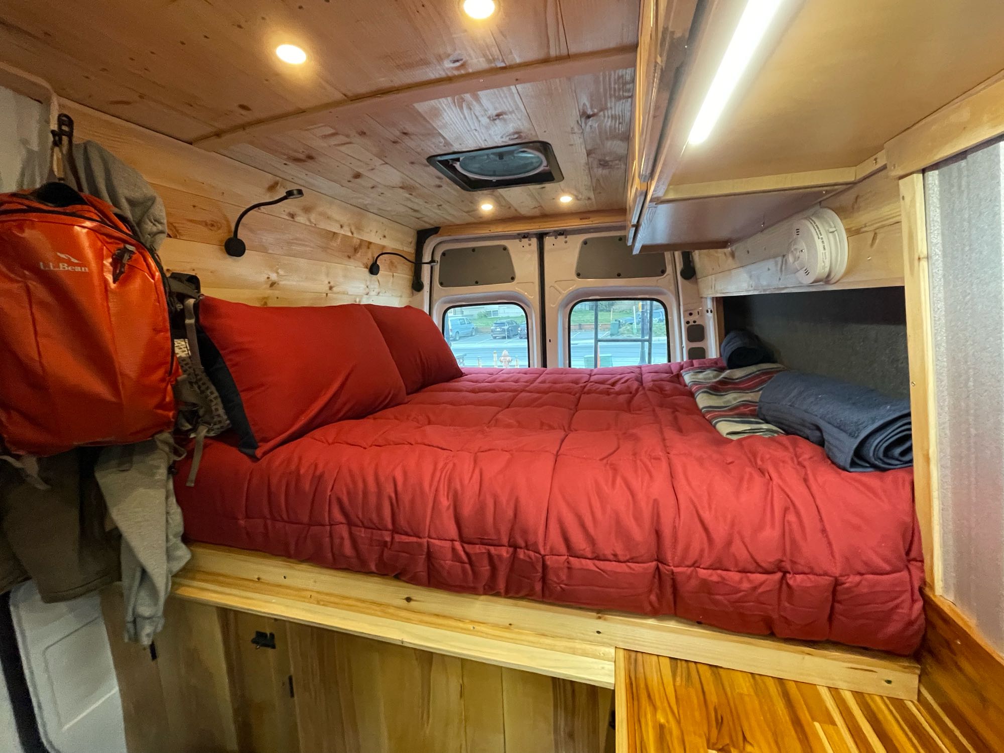 This rv rental features a cozy interior with a large red bed, warm wooden paneling, ceiling lights, and rear windows.. Mercedes Freightliner Sprinter 2021