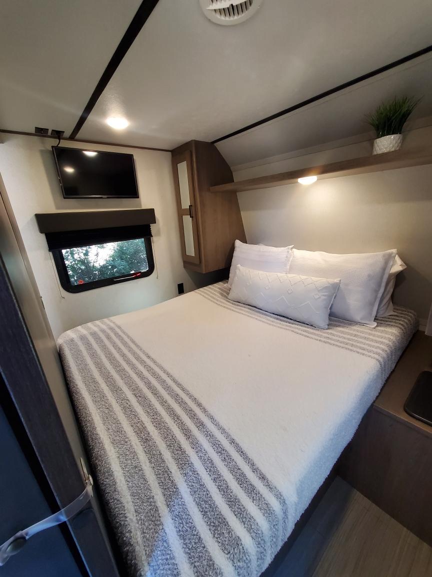 This cozy RV rental bedroom features a comfy bed, wall-mounted TV, window, and overhead storage.. Keystone RV Hideout Luxury 2021