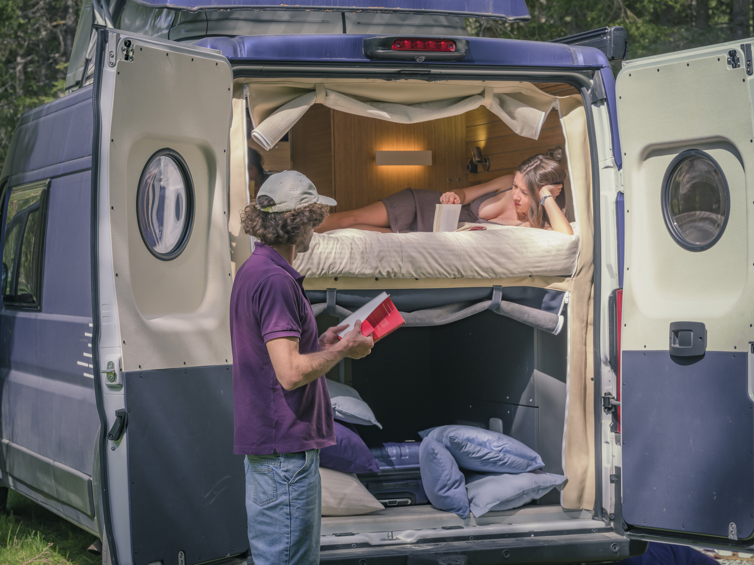 Relax in your RV rental! A man stands at the back of a campervan as a woman reads a book in the cozy bed inside.. Fiat Ducato MH2 2020