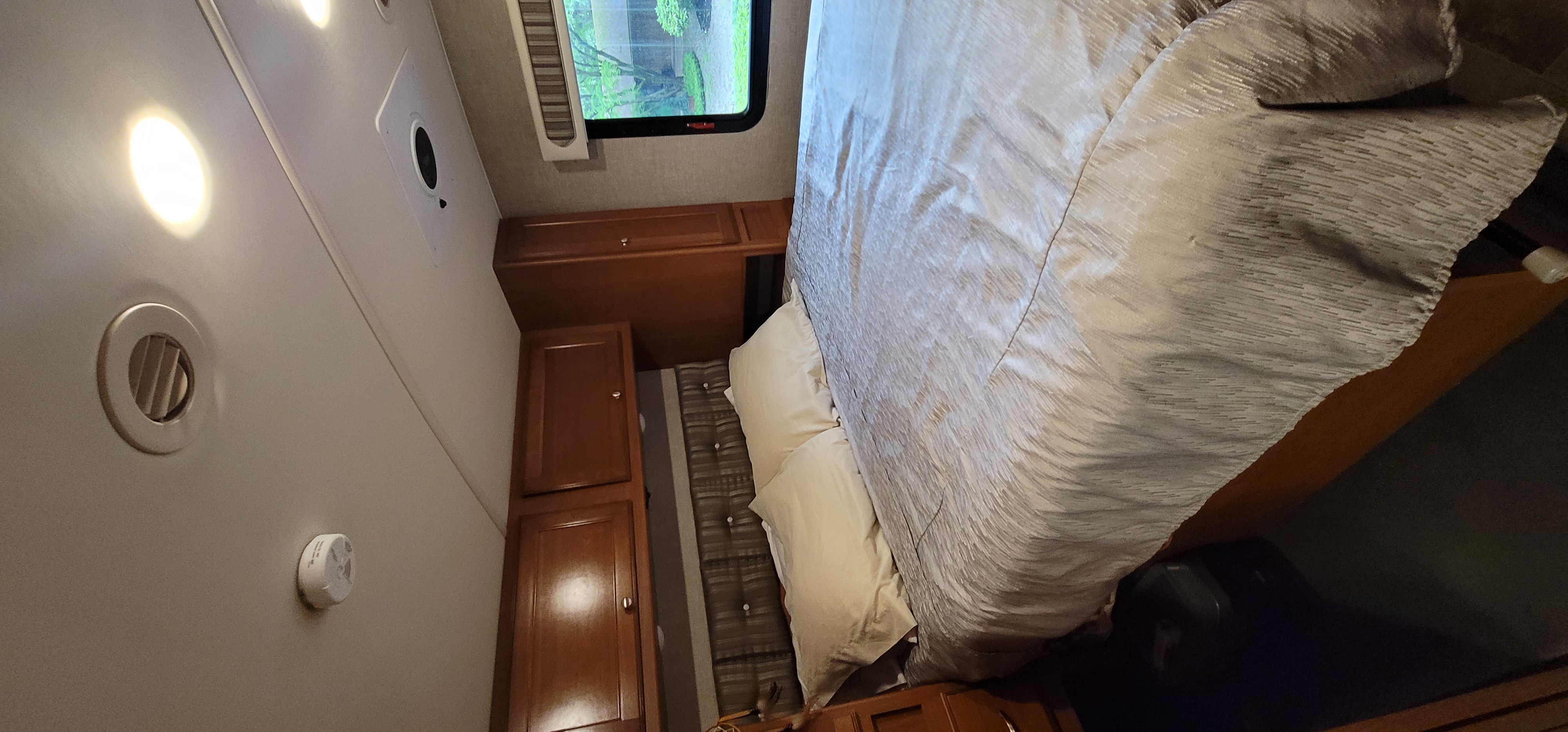 Inside an RV rental bedroom: A comfortable bed with light bedding, wooden cabinetry, window view, ceiling lights, and air vent.. Winnebago Vista 2019