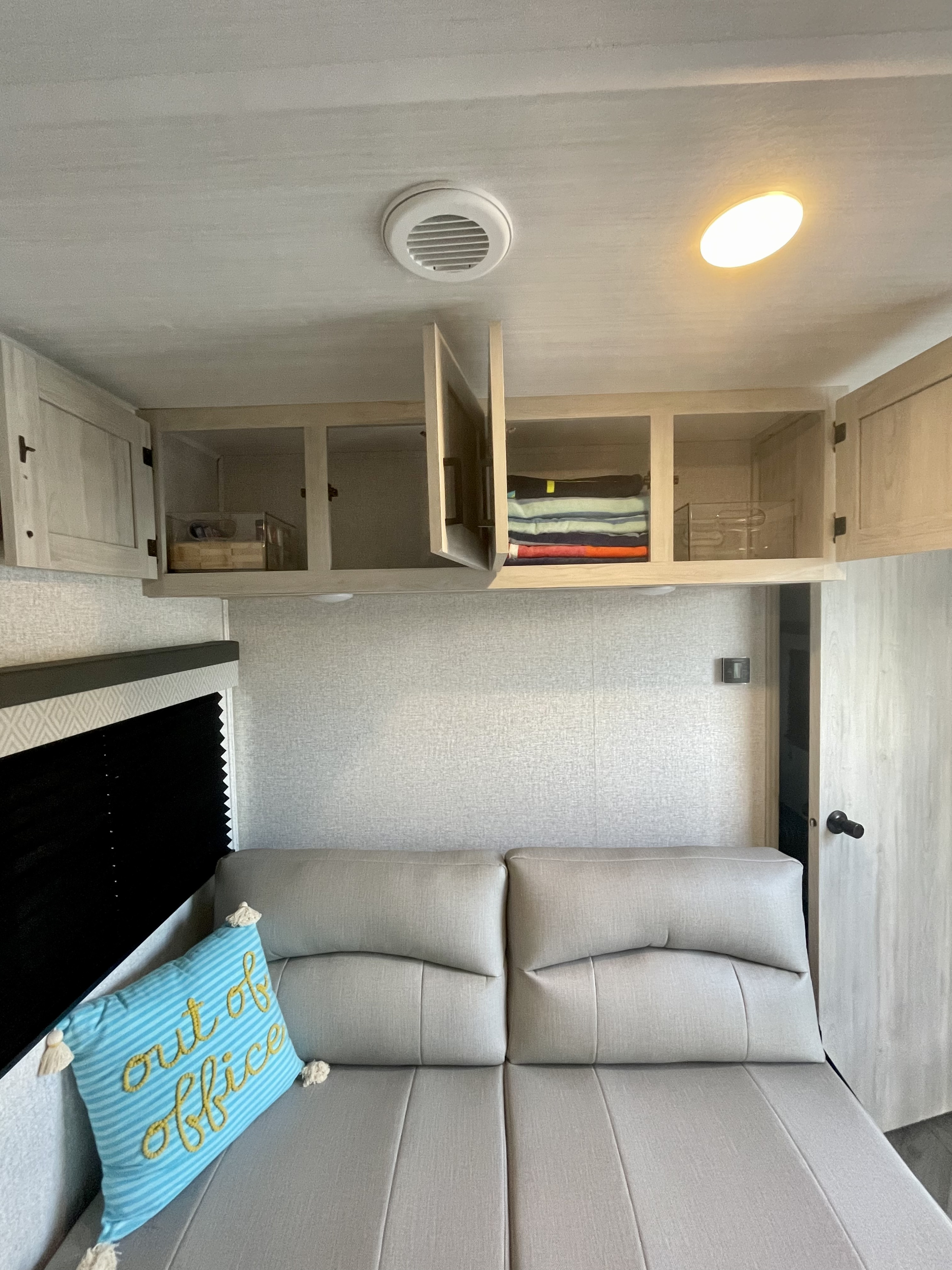 Cozy RV rental interior with opened overhead storage, a plush couch/bed, and an "out of office" throw pillow.. East to West Della Terra 2022