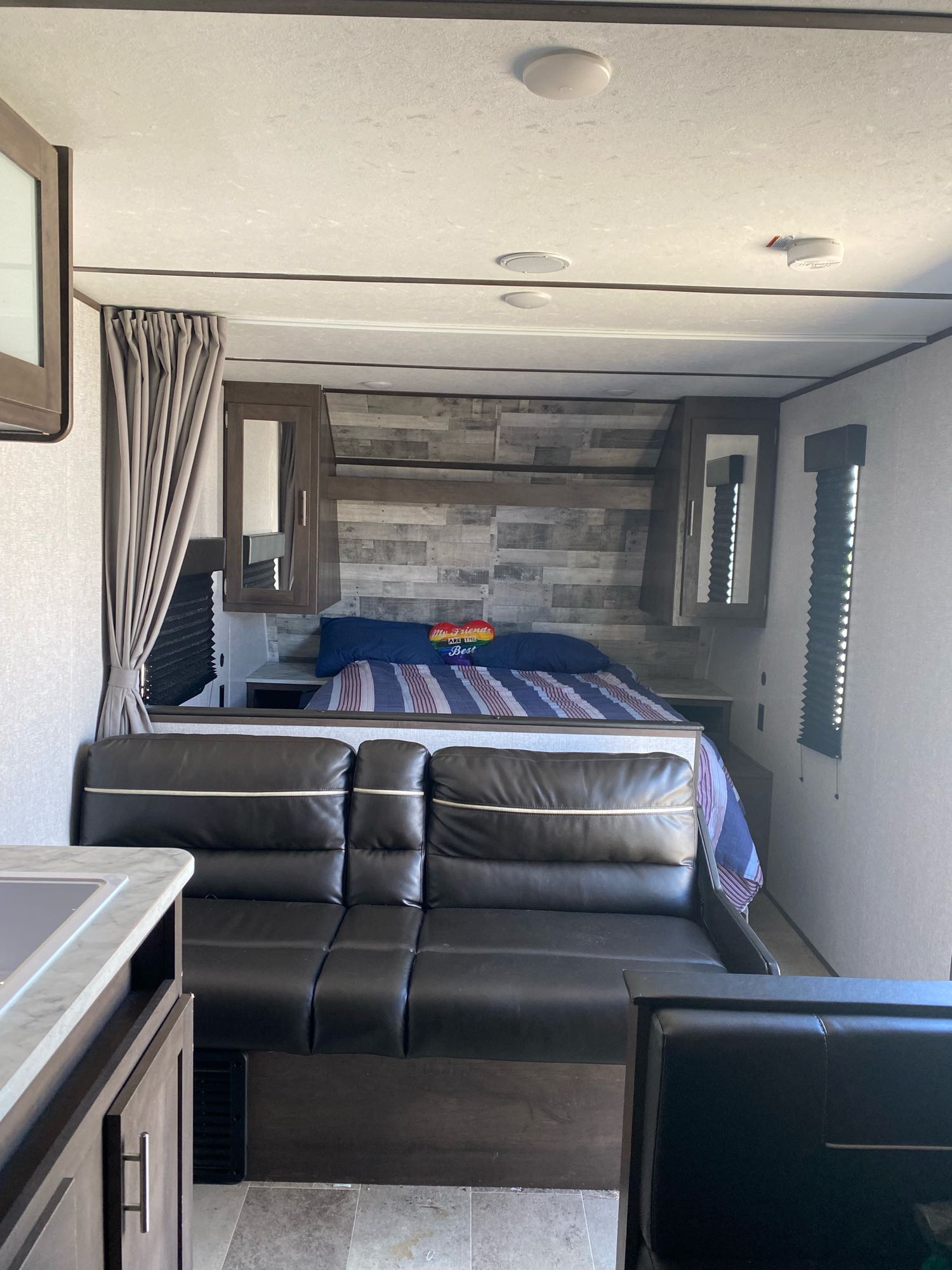 RV rental interior: A bedroom with a bed & grey wood wall, black sofa, and a kitchen counter visible on the left.. Dutchmen Coleman Lantern LT 2021