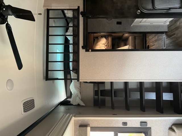 An RV rental interior featuring an elevated bunk bed with a safety rail, ceiling fan, and multi-level storage/shelving.. Jayco North Point 2021