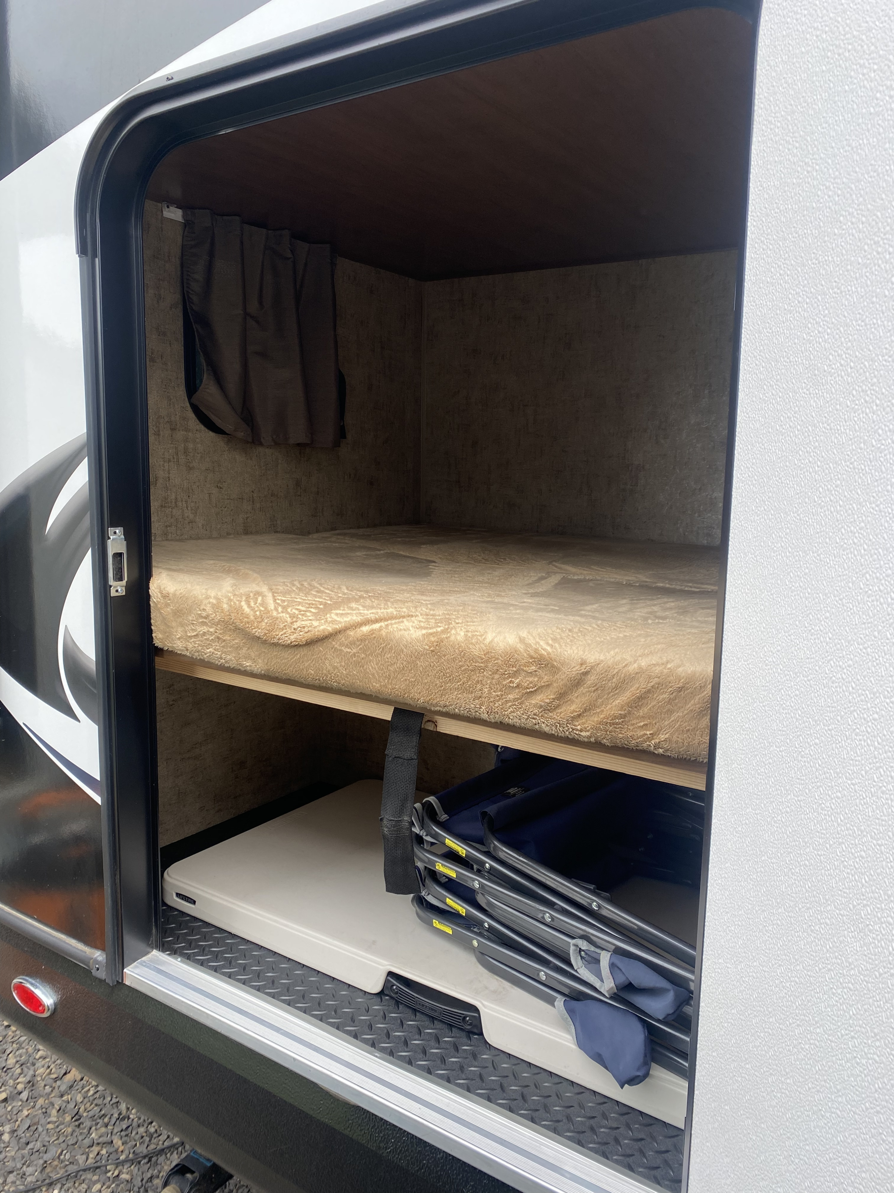 RV rental: Interior bunk with plush bedding and spacious lower storage holding a folding table and chairs, seen through an open hatch.. Outdoors Rv Manufacturing Creek Side 2021