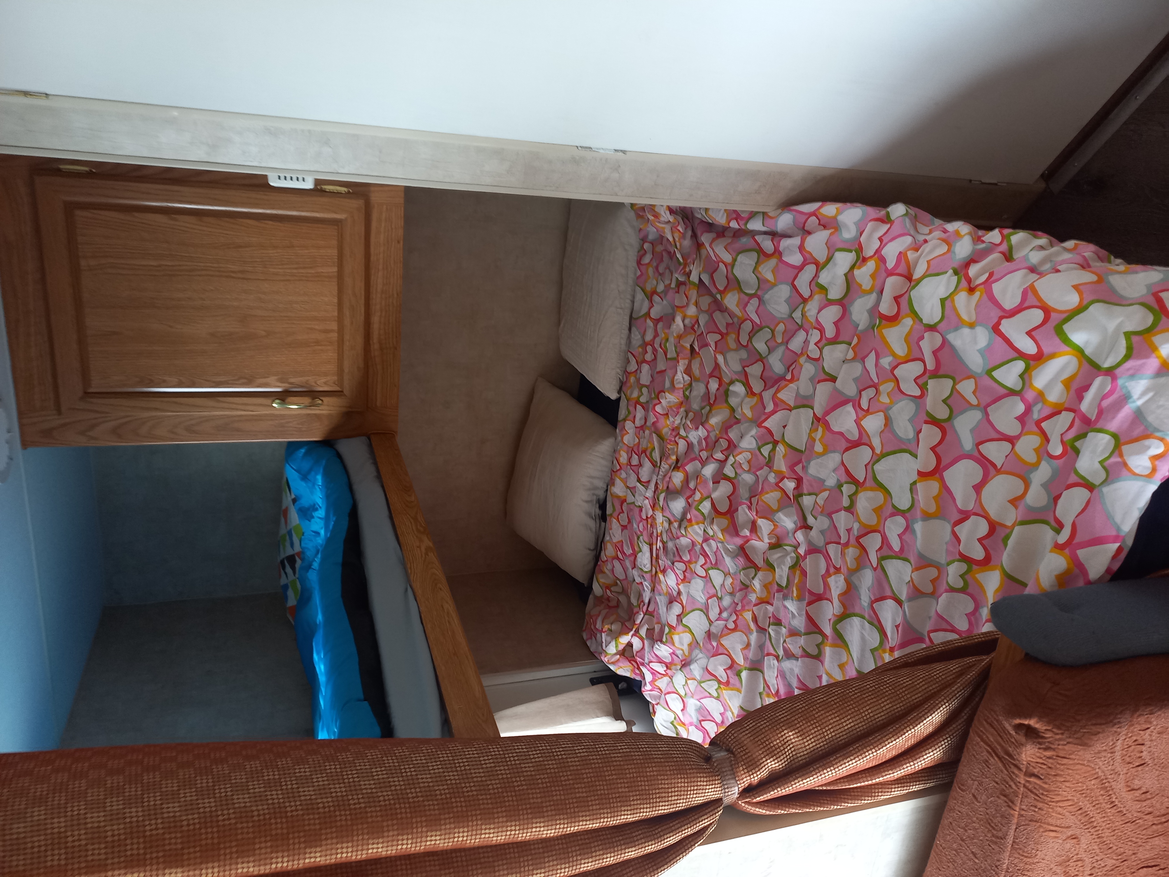 This RV rental interior features a cozy bed with heart-patterned bedding, adjacent to a wooden cabinet and lower bunk.. Fleetwood 240bh 2005