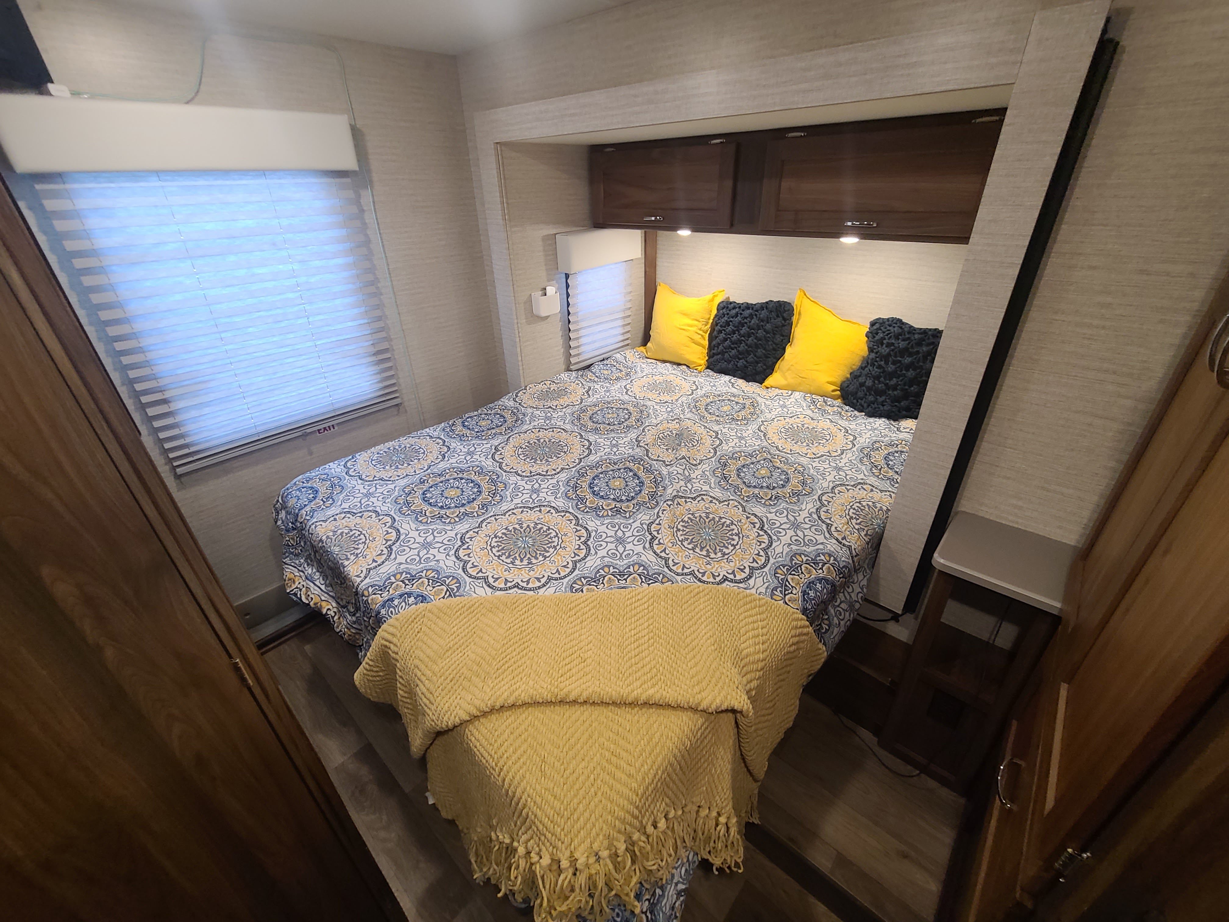Cozy RV rental bedroom with a mandala-patterned bedspread, yellow and textured gray pillows, and overhead wood cabinets.. Winnebago Outlook 2021