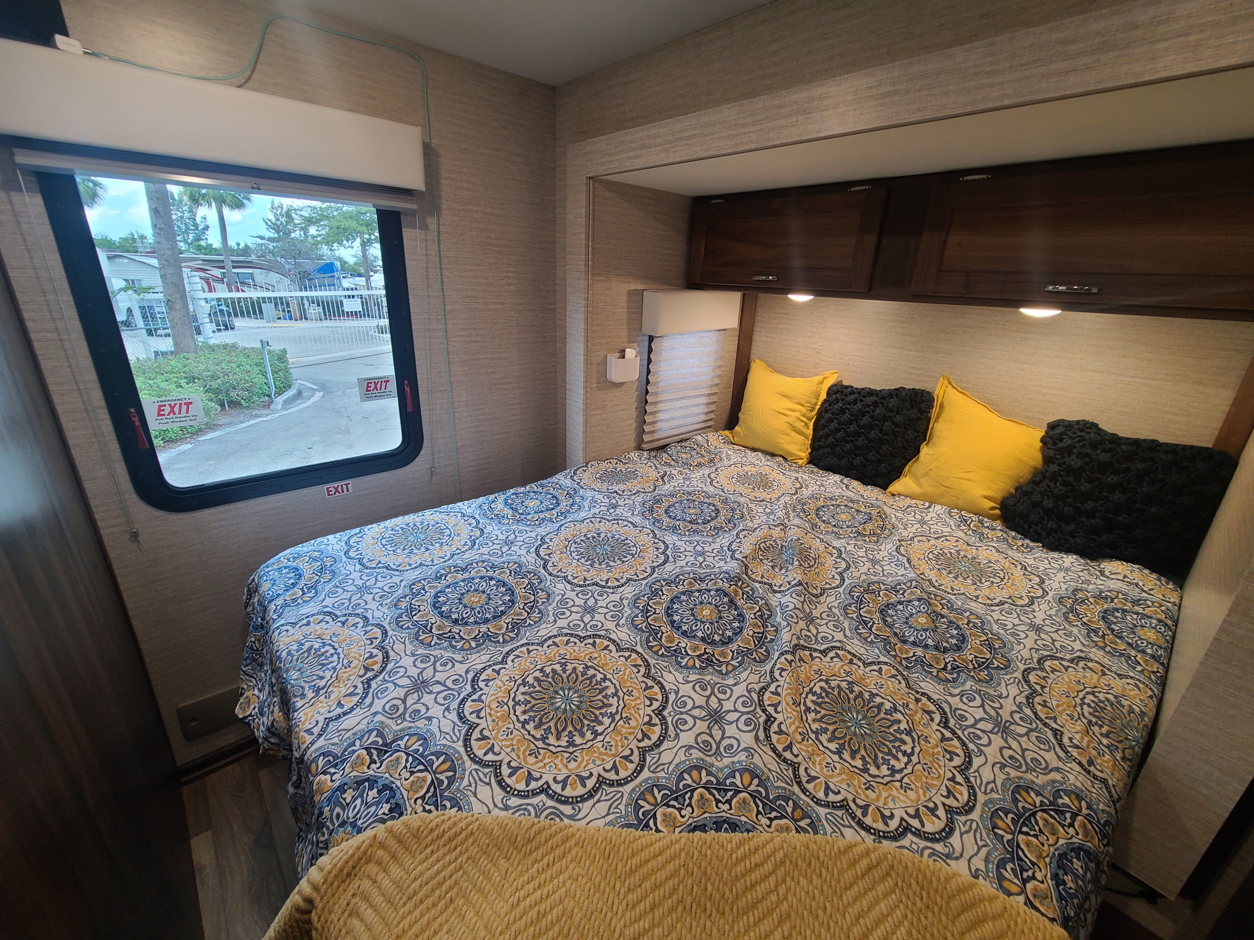 Inside an RV rental, a comfy bedroom with a vibrant patterned bed and sunny yellow pillows. Window offers a view of an RV park.. Winnebago Outlook 2021