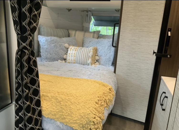 Experience comfort in this RV rental's cozy bedroom, featuring fluffy grey bedding, a yellow throw, and nature views.. Airstream Airstream 2020