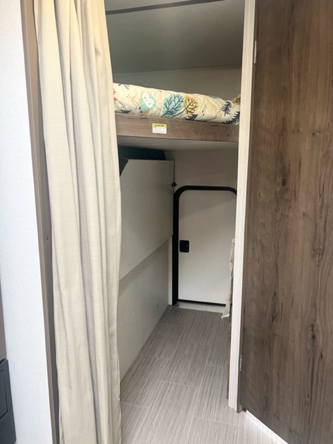 Cozy bunk bed area in an RV rental, featuring a colorful duvet, a light curtain, and a tiled hallway leading to a door.. Keystone RV Passport SL 2020