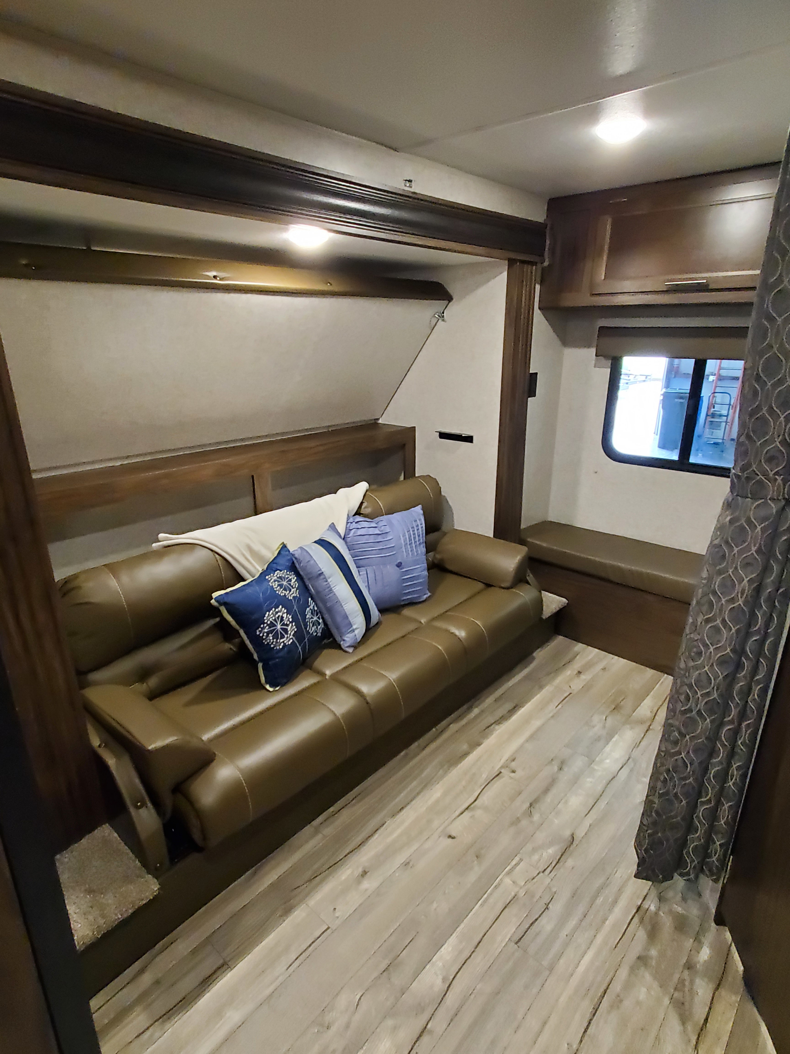 RV rental interior with a brown leatherette sofa, blue pillows, and an empty bunk bed. Features wood-look flooring & cabinetry.. Forest River Cherokee Grey Wolf 2019
