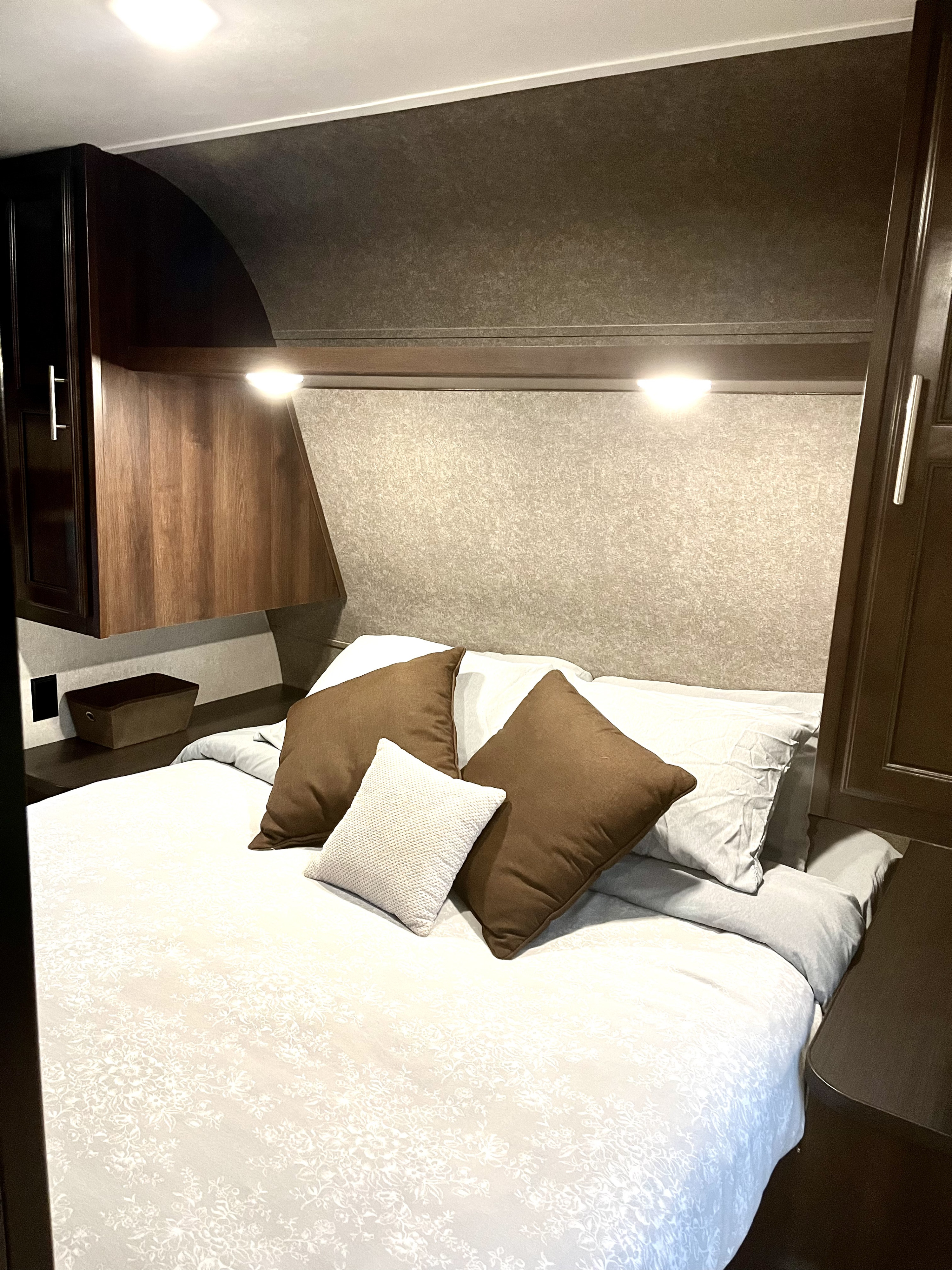 Cozy RV rental bedroom with a neat bed, white patterned bedding, brown pillows, and dark wood overhead cabinets.. Jayco Jay Flight SLX Rocky Mtn Edition 2019