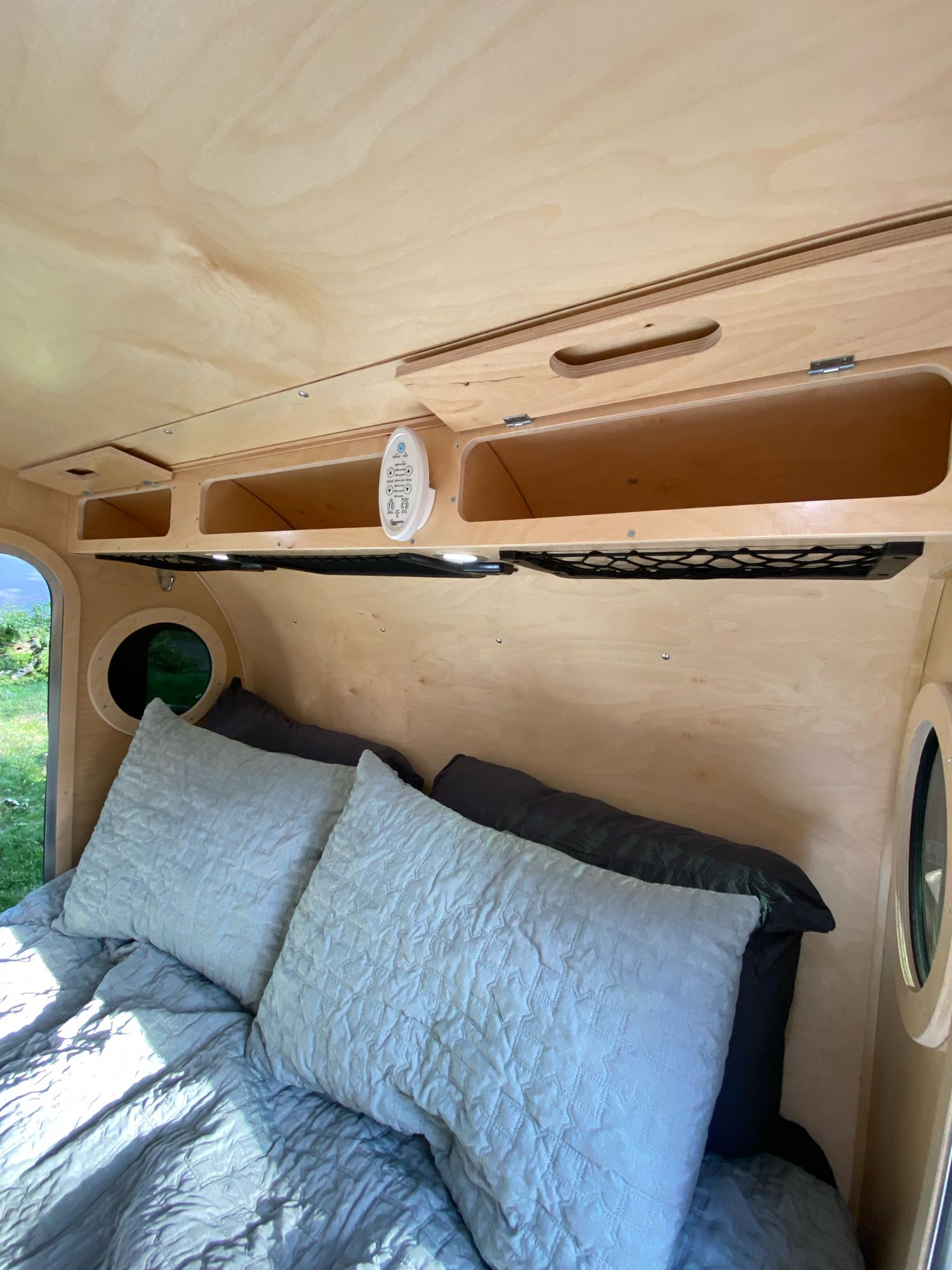 Compact RV rental bedroom with light wood paneling, cozy bed, overhead storage, and control panel.. Teardrops Northwest Trek 2022