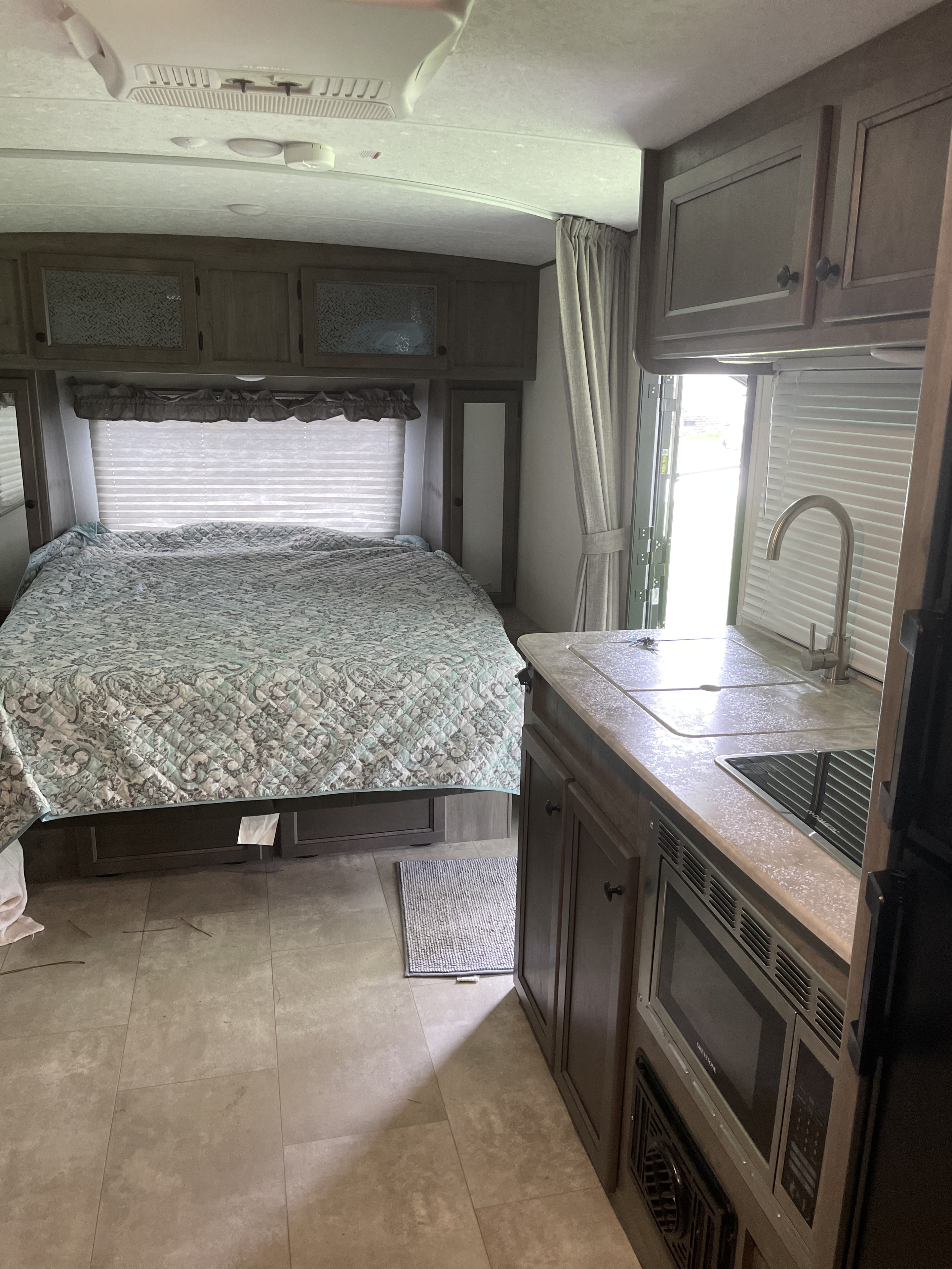 Inside an RV rental, showcasing a comfortable bed, overhead storage, and a compact kitchenette with a sink, microwave, and outdoor access.. Coachmen Apex Nano 2021