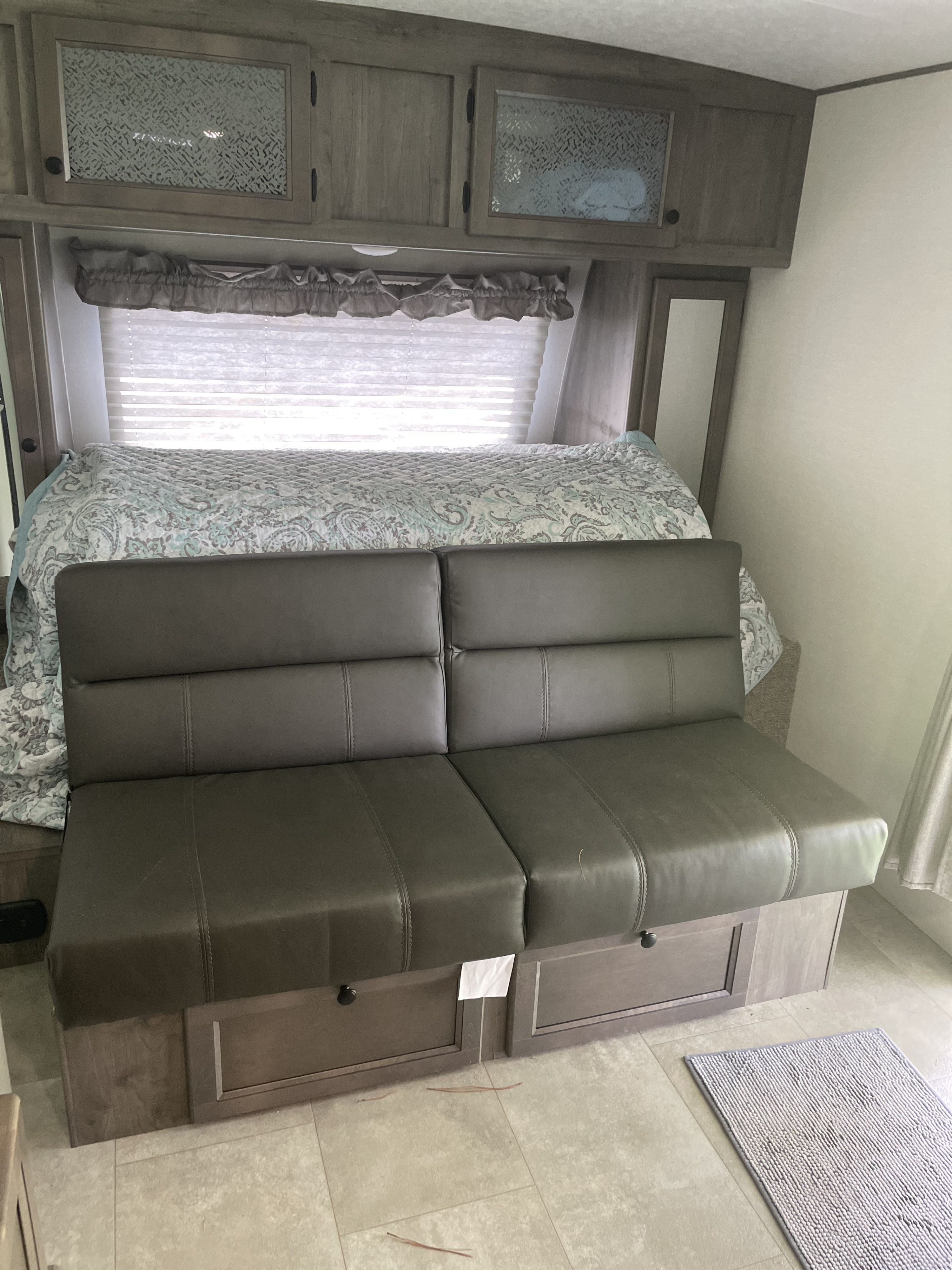 View inside an RV rental: a made bed, grey sofa with storage drawers, overhead cabinets, and window.. Coachmen Apex Nano 2021