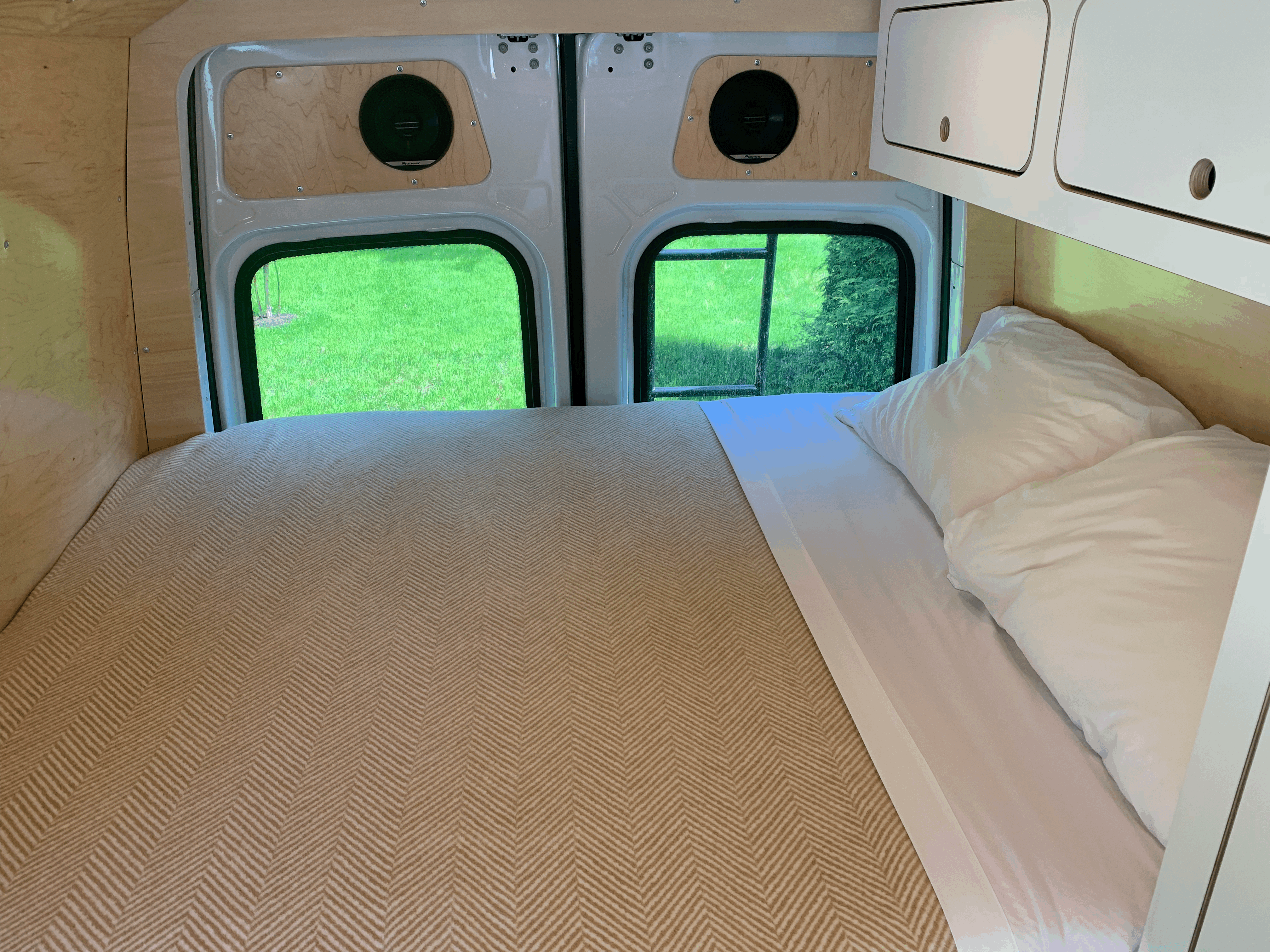 Cozy RV rental interior: a bed with herringbone blanket, wood walls, overhead storage, & views of nature through rear windows.. Mercedes-Benz Sprinter 2021