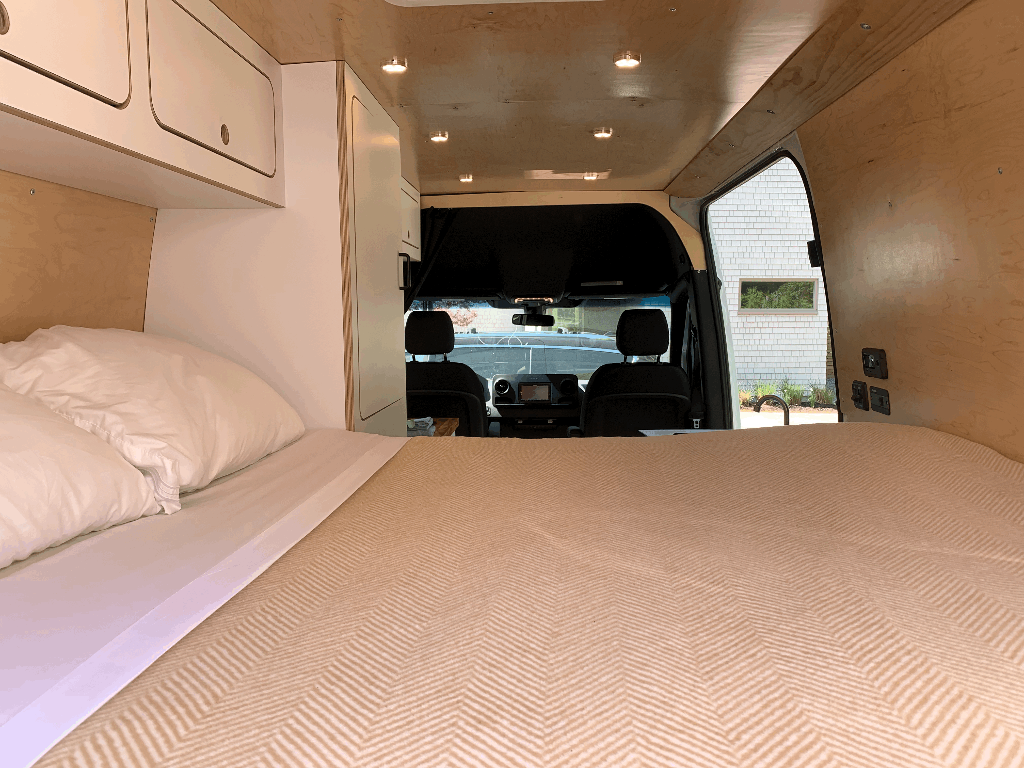 Cozy RV rental interior featuring a comfortable bed, overhead cabinets, warm wooden paneling, and the driver's cab.. Mercedes-Benz Sprinter 2021