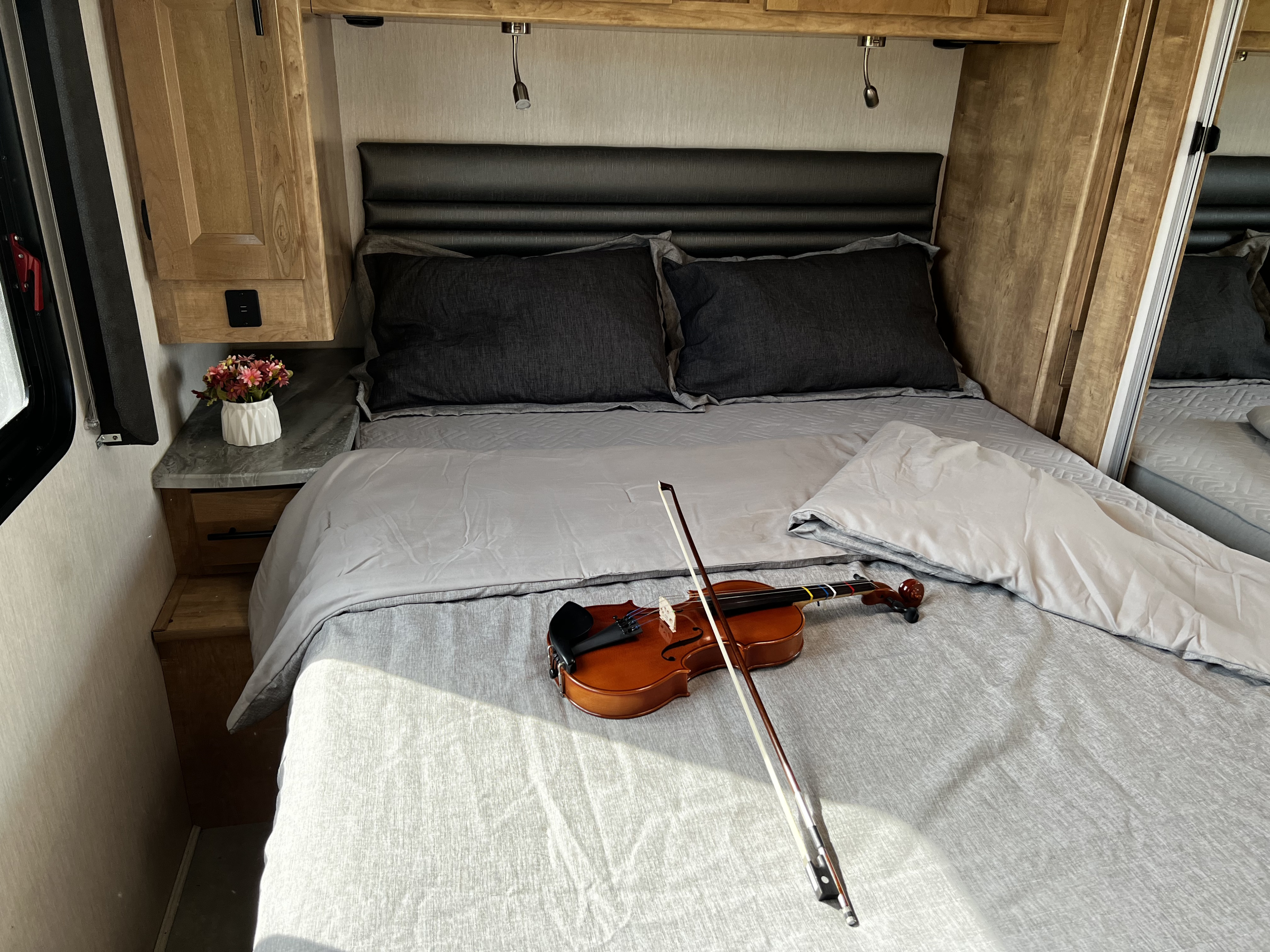 Cozy RV rental bedroom with grey bedding, a black headboard, wooden cabinets, and a violin on the bed.. Coachmen Leprechaun 2021